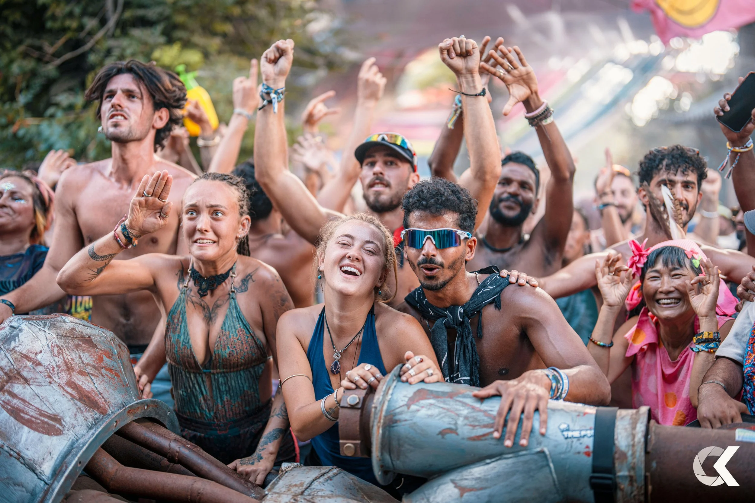 A diverse group of people enjoying a lively outdoor music festival, dancing and smiling, some shirtless, with trees and a tent in the background.