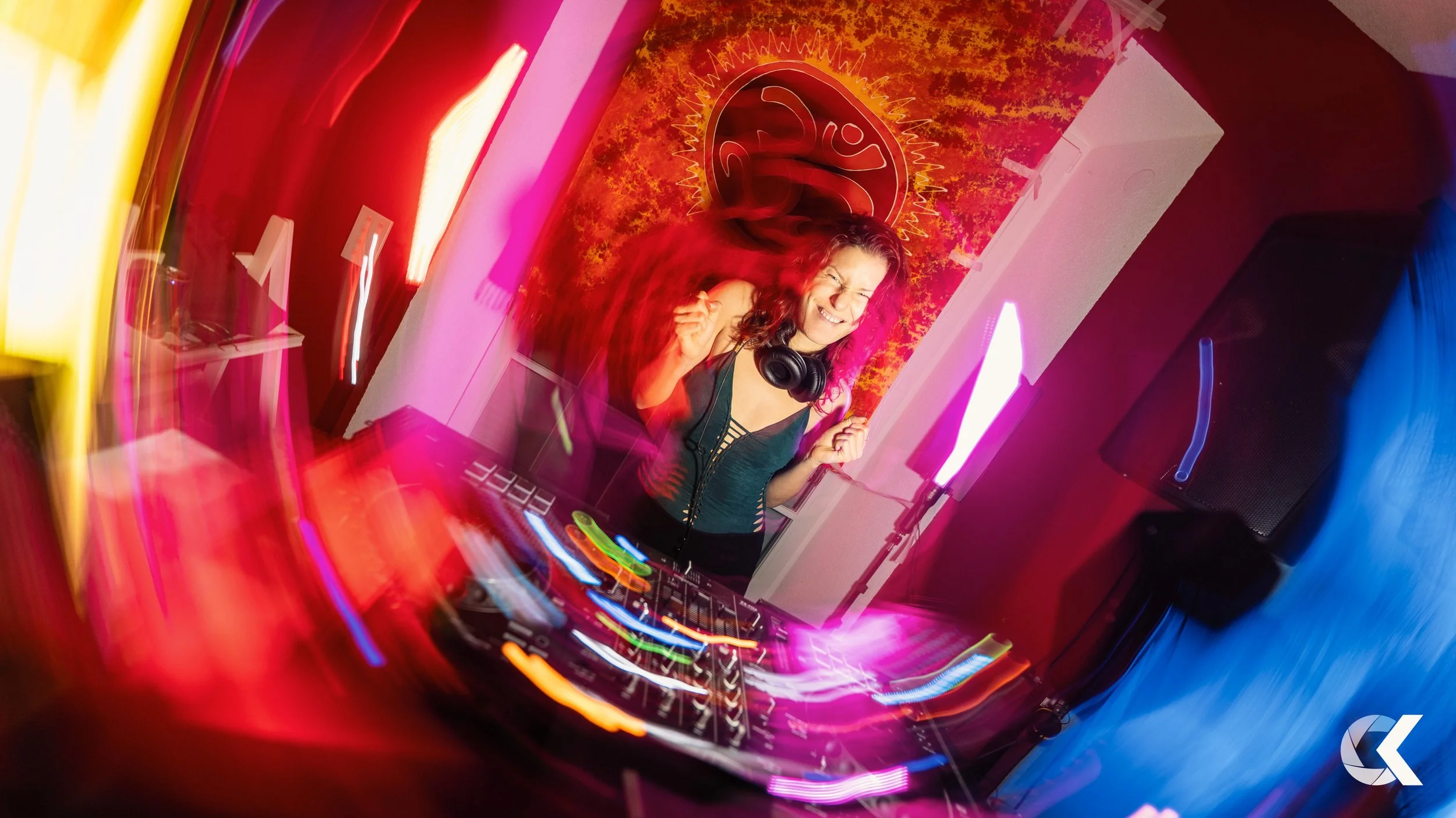 A female DJ with headphones around her neck dancing and smiling at her DJ booth with colorful lights and a vibrant red and orange background.
