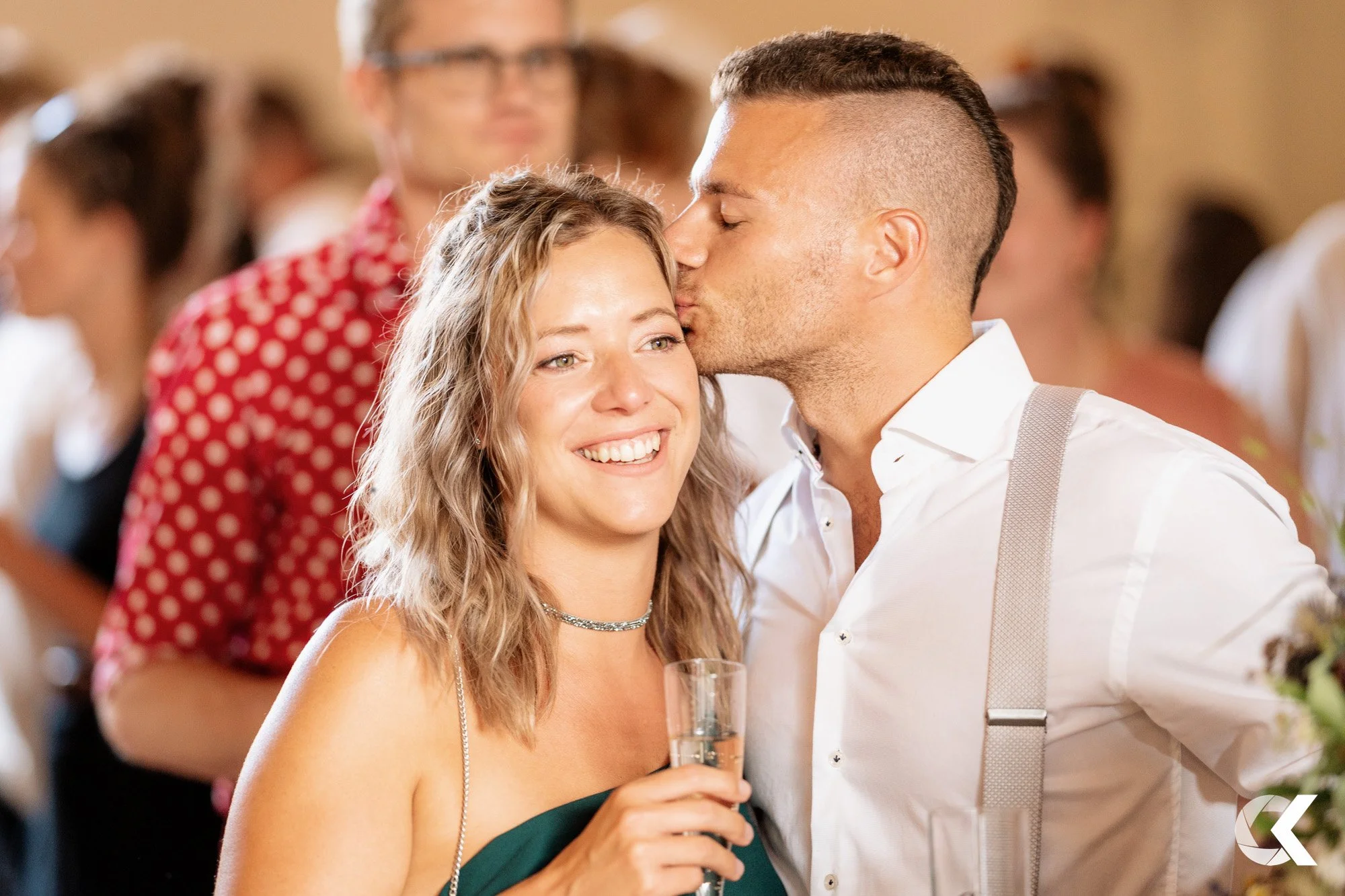 A man kissing a woman on the forehead at a celebration or wedding reception while holding a glass of champagne. The woman is smiling, holding a drink, and wearing a strapless dark green dress and a choker necklace, with other guests blurred in the ba