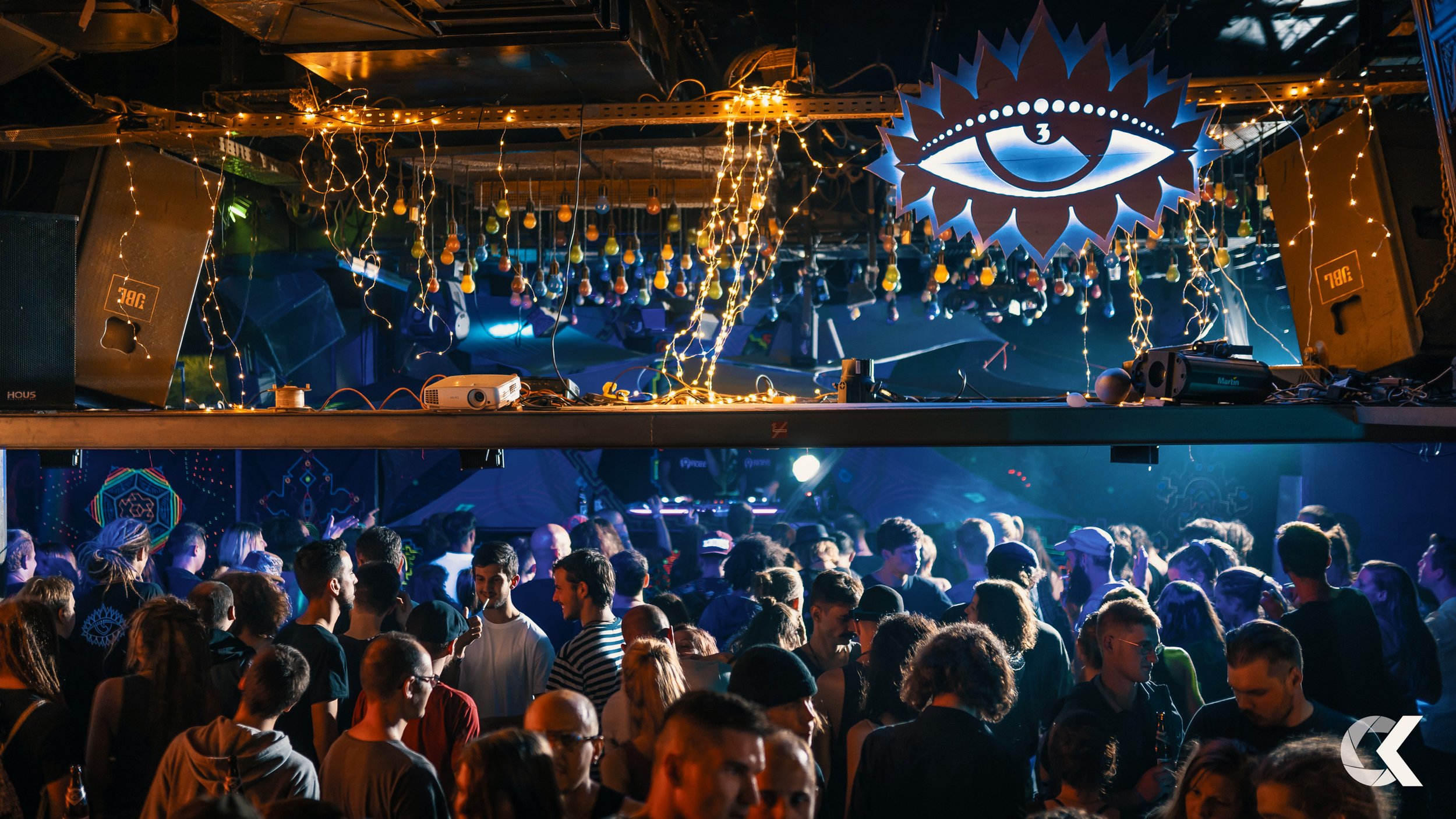 Crowd of people at a nightclub dance floor with colorful lighting and decorations, and a DJ booth with lights and equipment overhead.