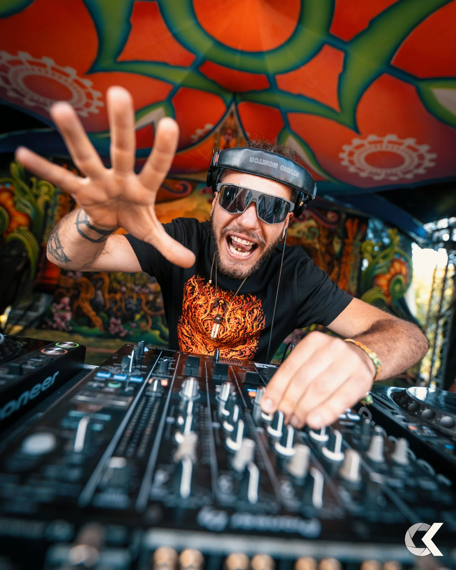 DJ wearing sunglasses and headphones, reaching towards the camera while performing at a vibrant, colorful outdoor event with psychedelic art backdrop.
