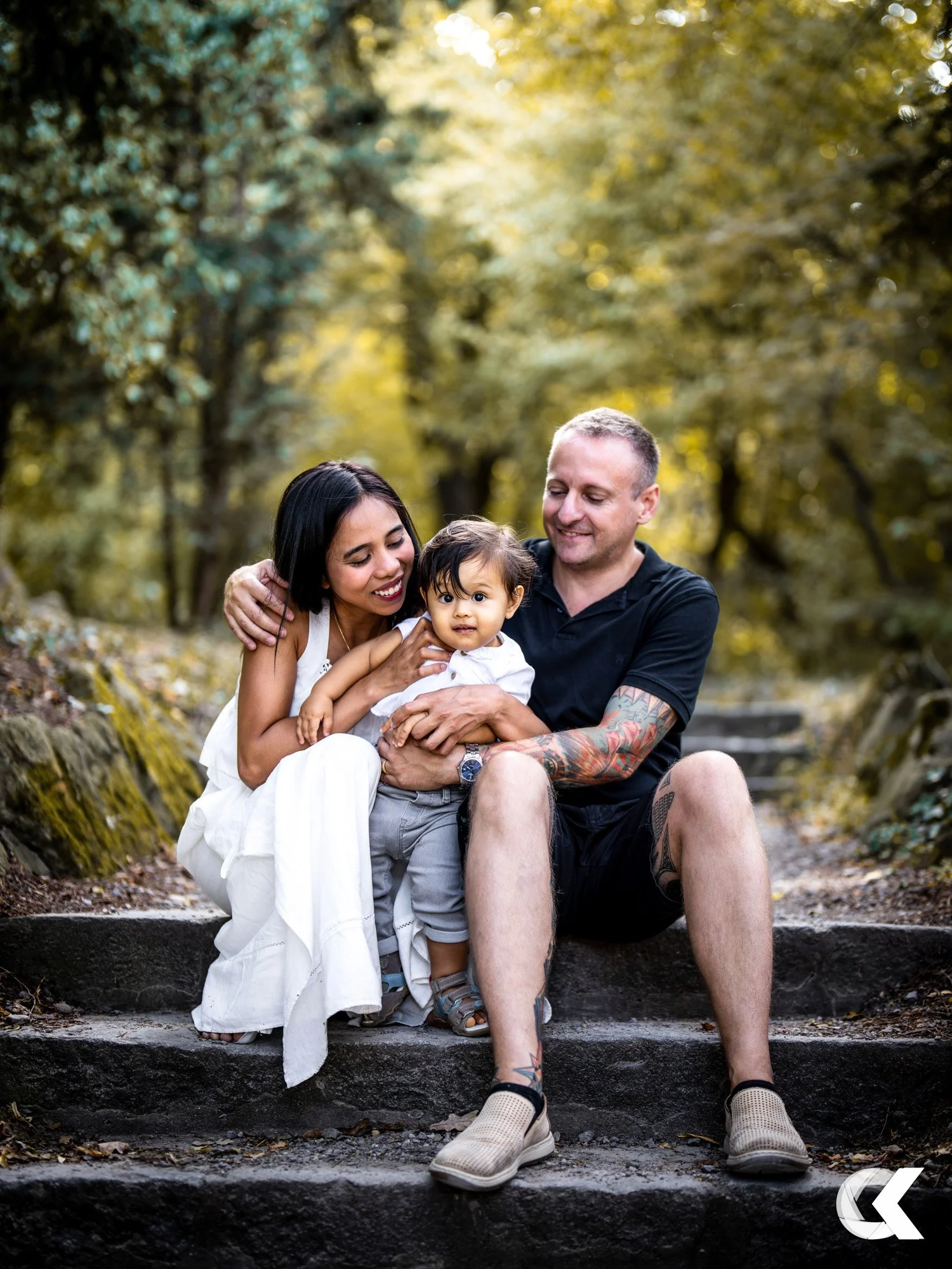 A family of three sitting on outdoor steps in a wooded area, smiling and hugging, with sunlight filtering through trees in the background.