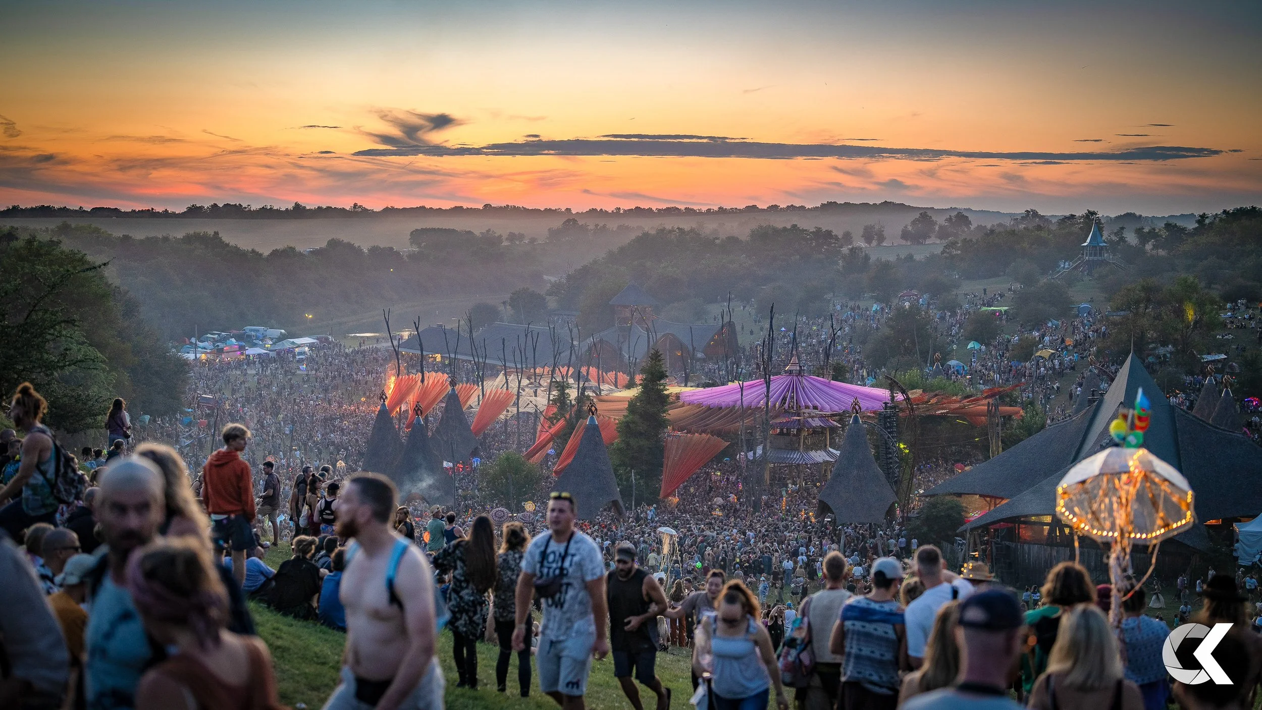 Crowd gathering at an outdoor music festival during sunset with colorful tents and trees in the background.