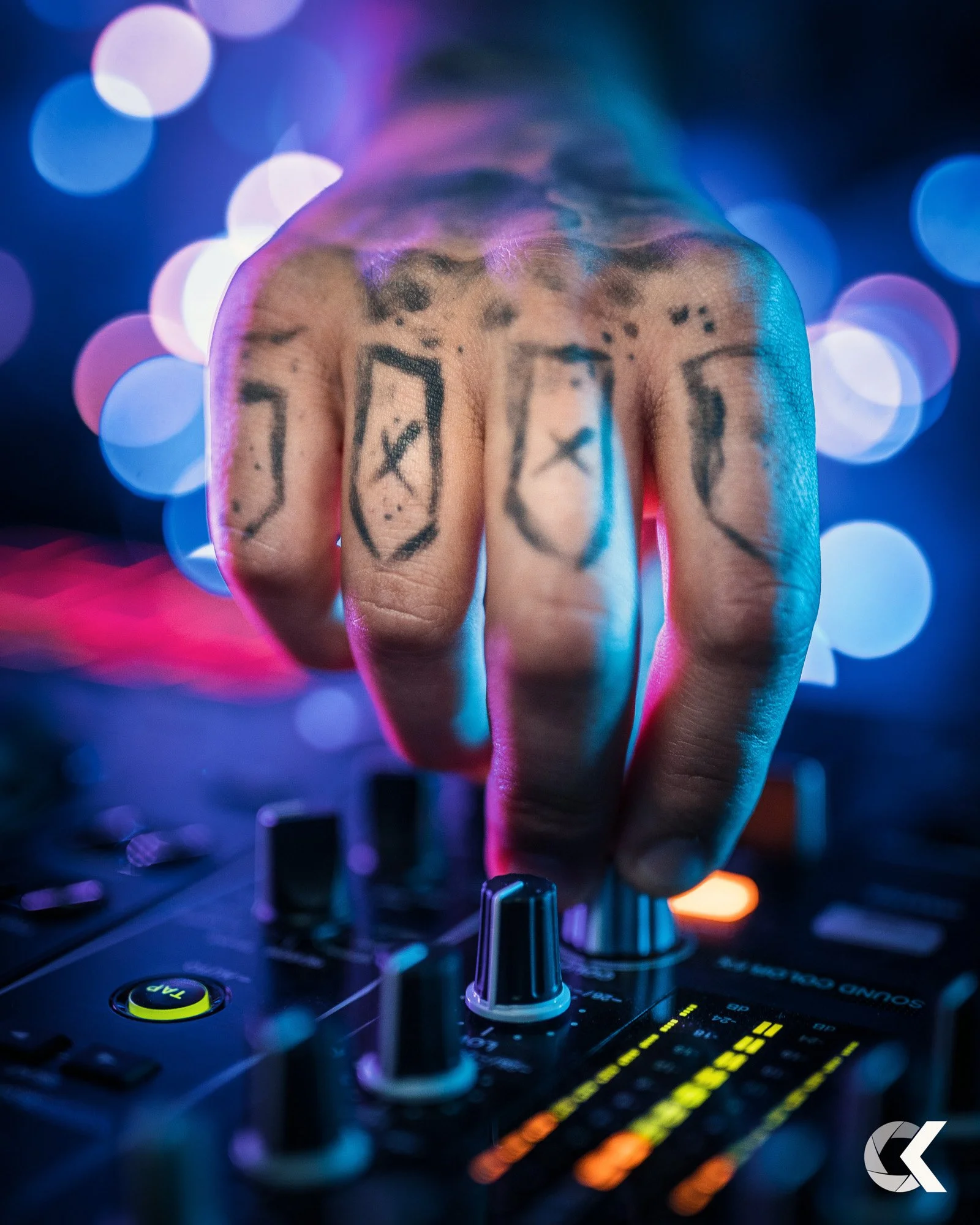 Close-up of a hand with tattoos on fingers, adjusting audio mixer controls with colorful bokeh background.