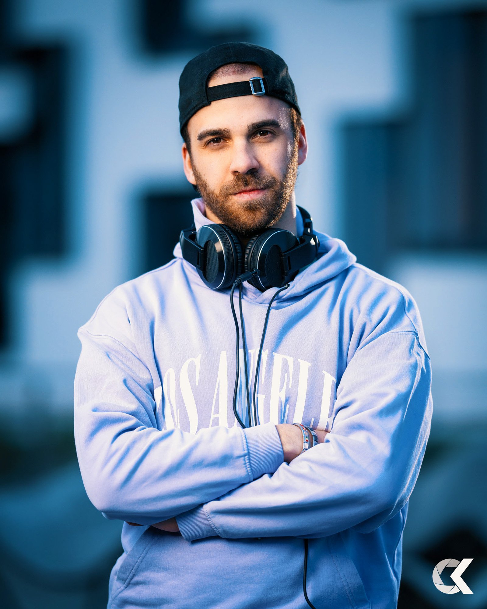 Young man with a beard wearing a black snapback hat backwards, headphones around his neck, a light purple hoodie with 'Los Angeles' printed on it, and bracelets on his wrist, standing outdoors with arms crossed, facing camera.