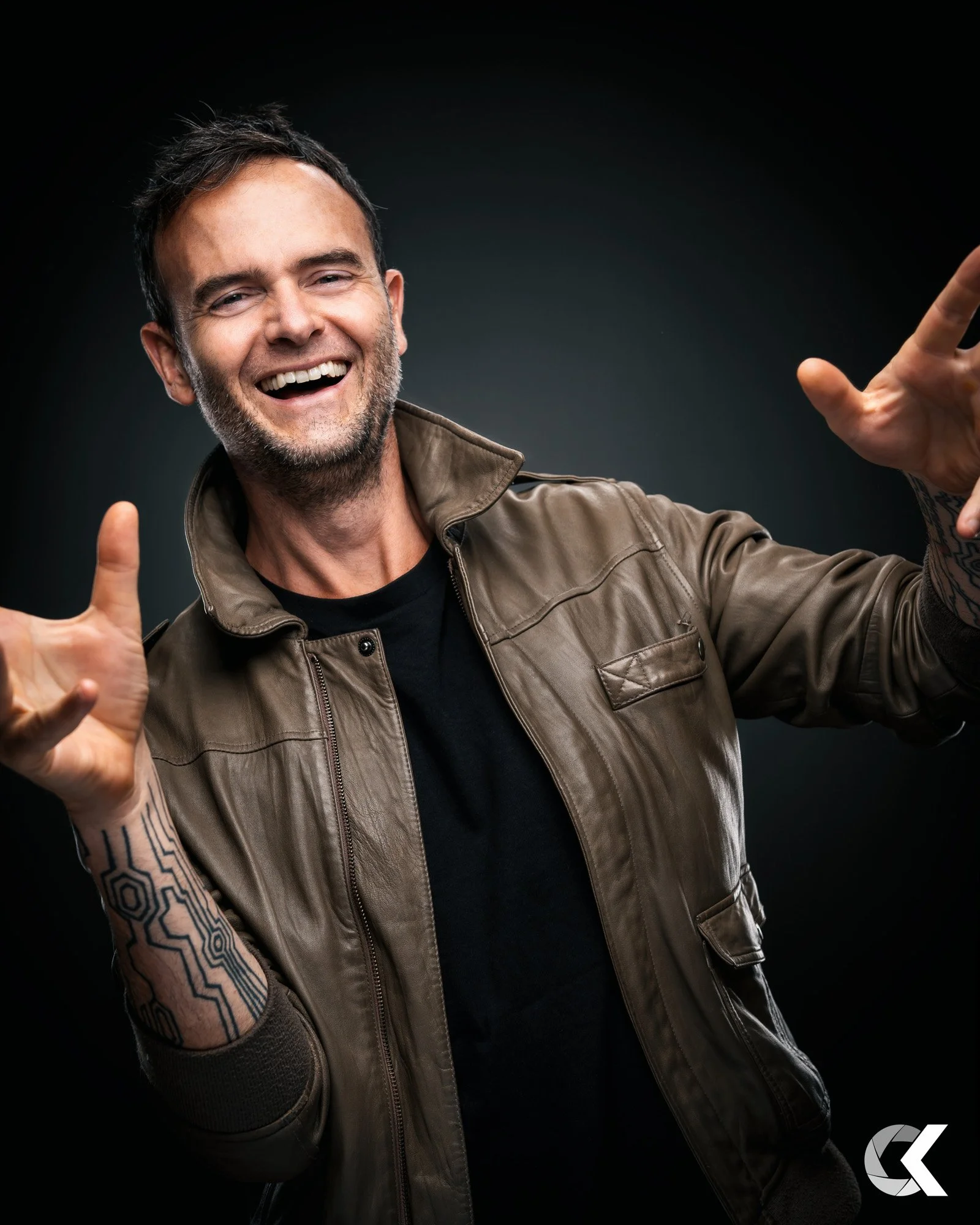 A smiling man with dark hair and a beard, wearing a brown leather jacket and black shirt, gesturing with both hands in front of a dark background.