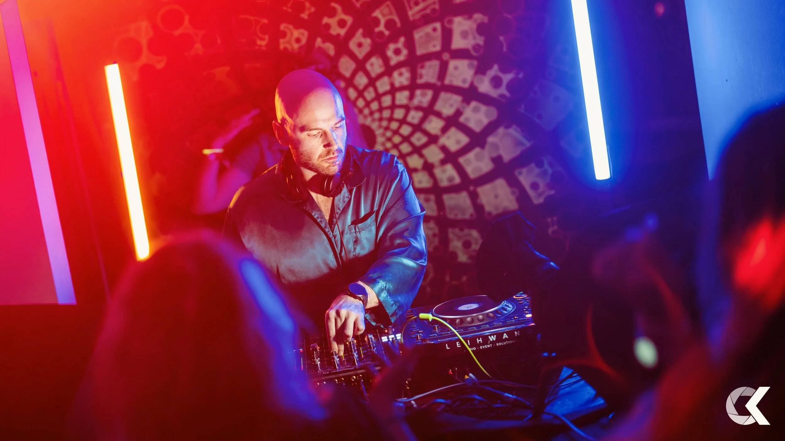 A DJ with a shaved head and beard wearing headphones around his neck, operating a DJ mixer in a nightclub with colorful lighting and a patterned wall in the background.