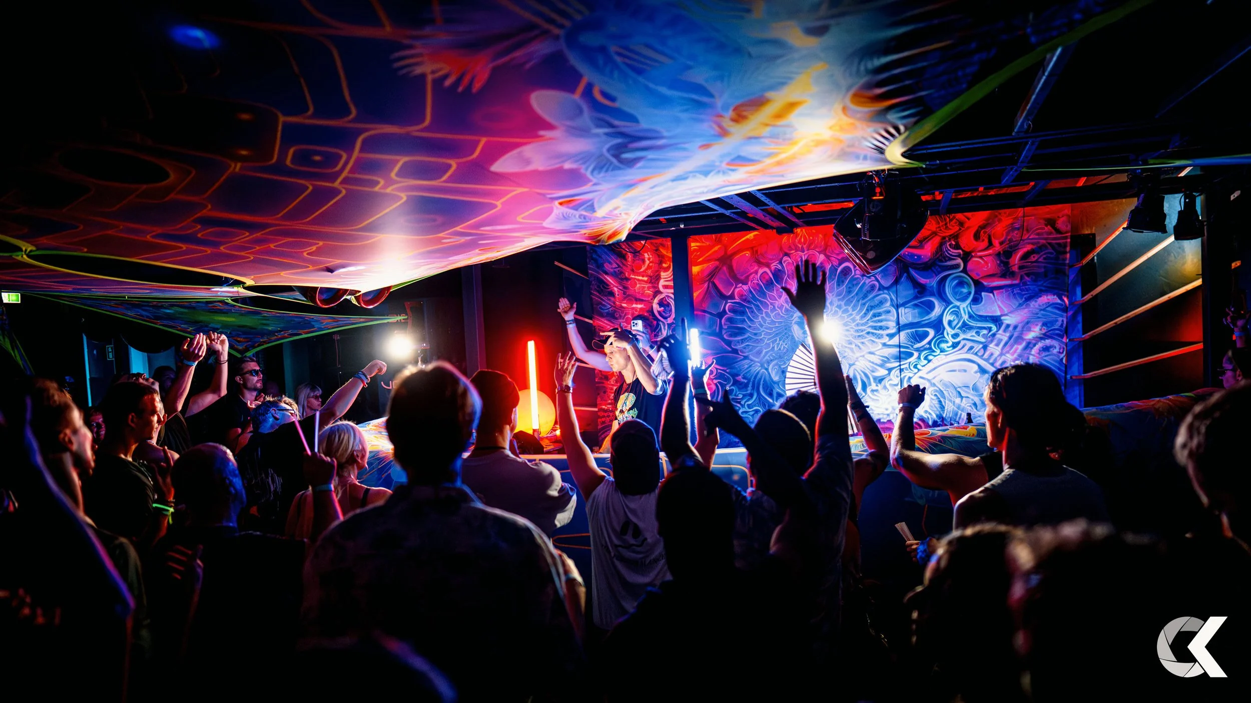 A group of people dancing and enjoying music at a vibrant, colorful nightclub with psychedelic art on the walls and ceiling, illuminated by neon lights.