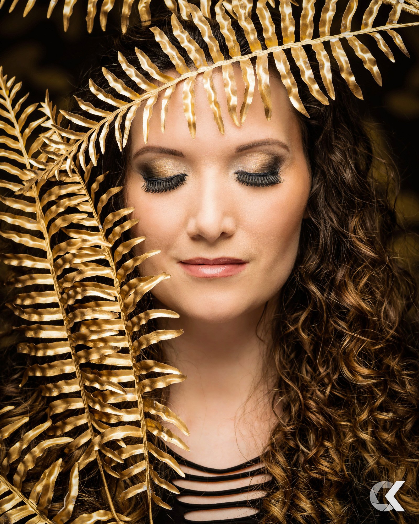 A woman with curly hair and gold eyeshadow has her eyes closed and is surrounded by gold fern leaves.