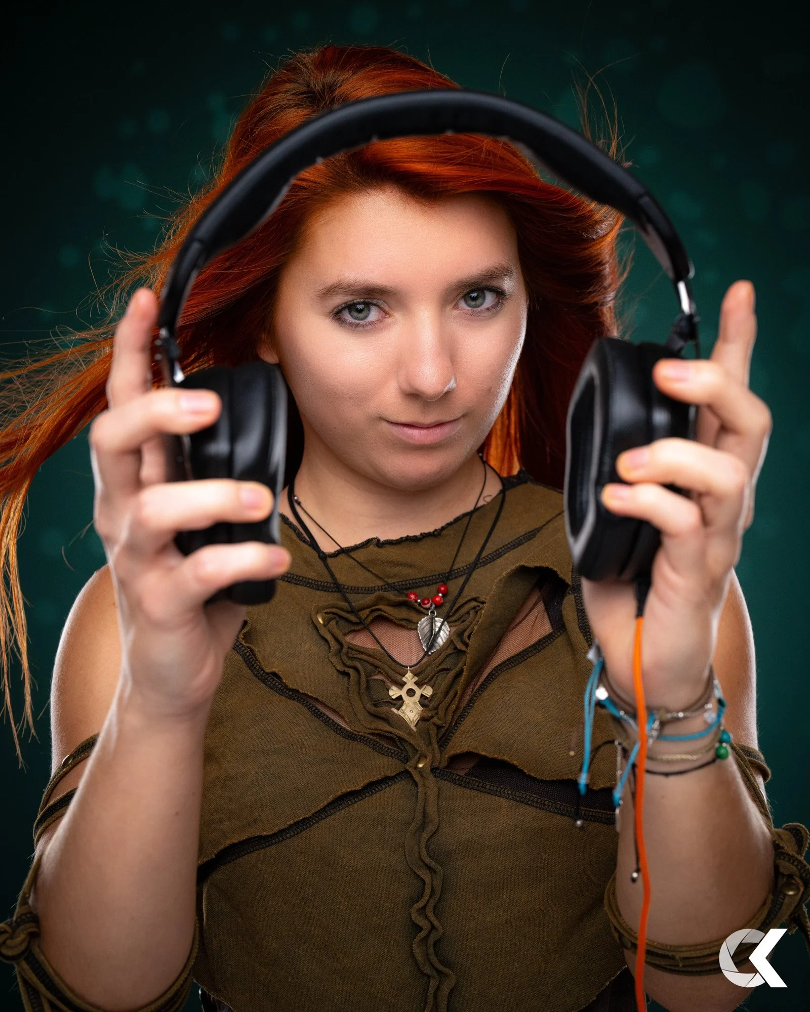 Junge Frau mit roten Haaren hält schwarze Kopfhörer vor ihrem Gesicht, schaut direkt in die Kamera, trägt braunes Top und mehrere Halsketten, bunte Armbänder, vor dunklem, unscharfem Hintergrund.