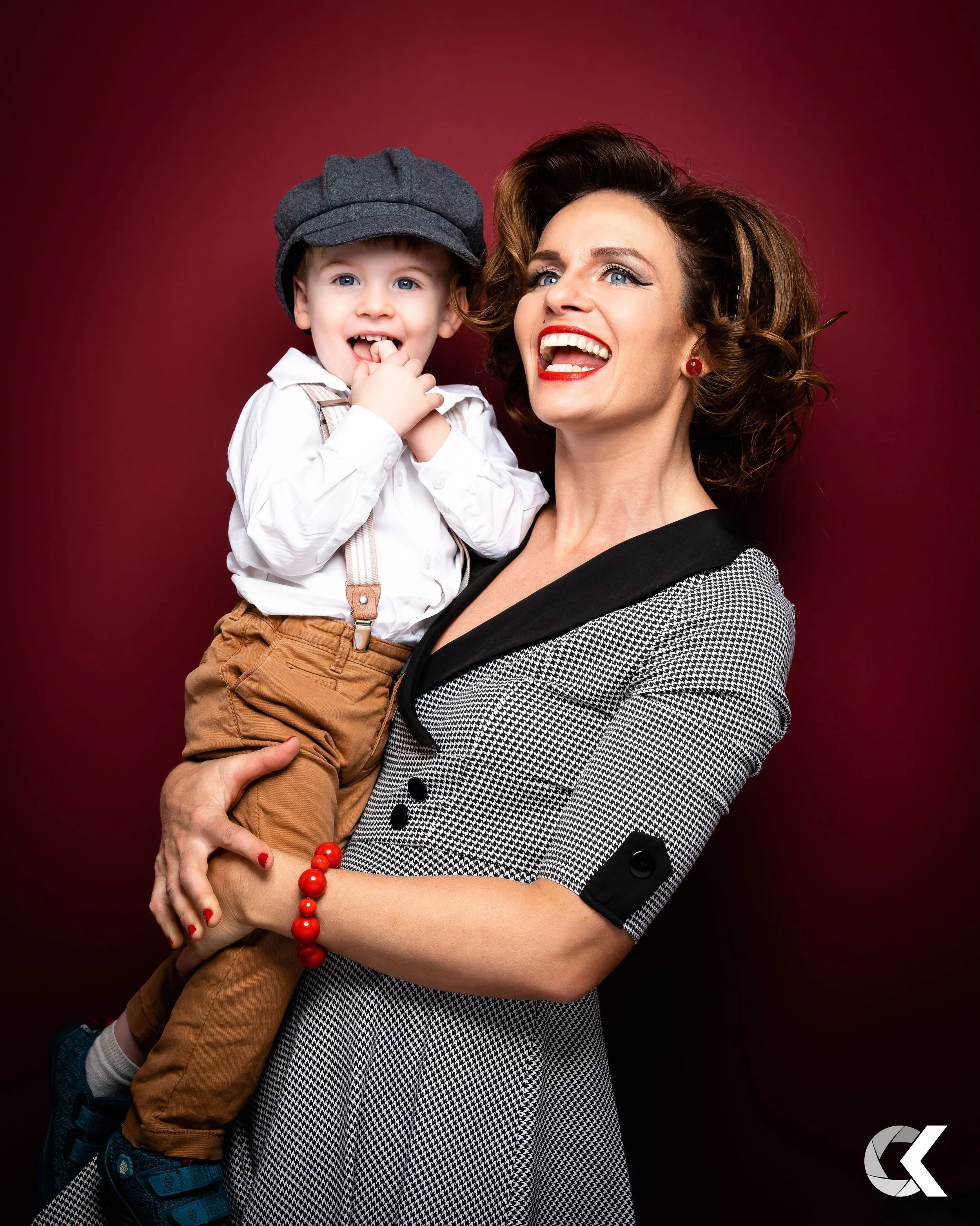 A woman with curly hair, red lipstick, and a black-and-white dress holding a young boy with a gray hat, white shirt, and tan pants on a red background.