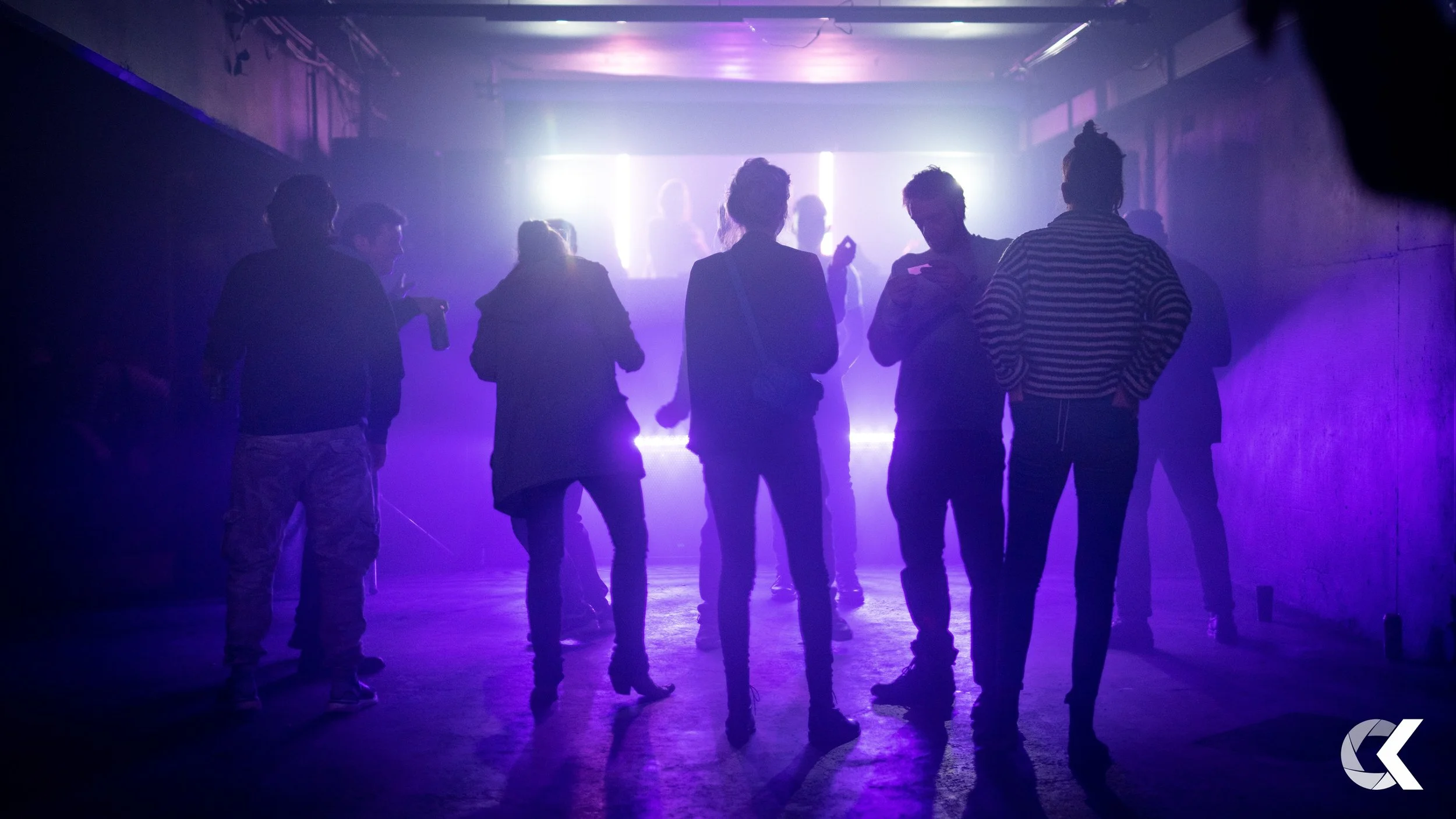 Group of people standing in a dimly lit indoor nightclub or concert venue, silhouetted against bright purple and white stage lights.