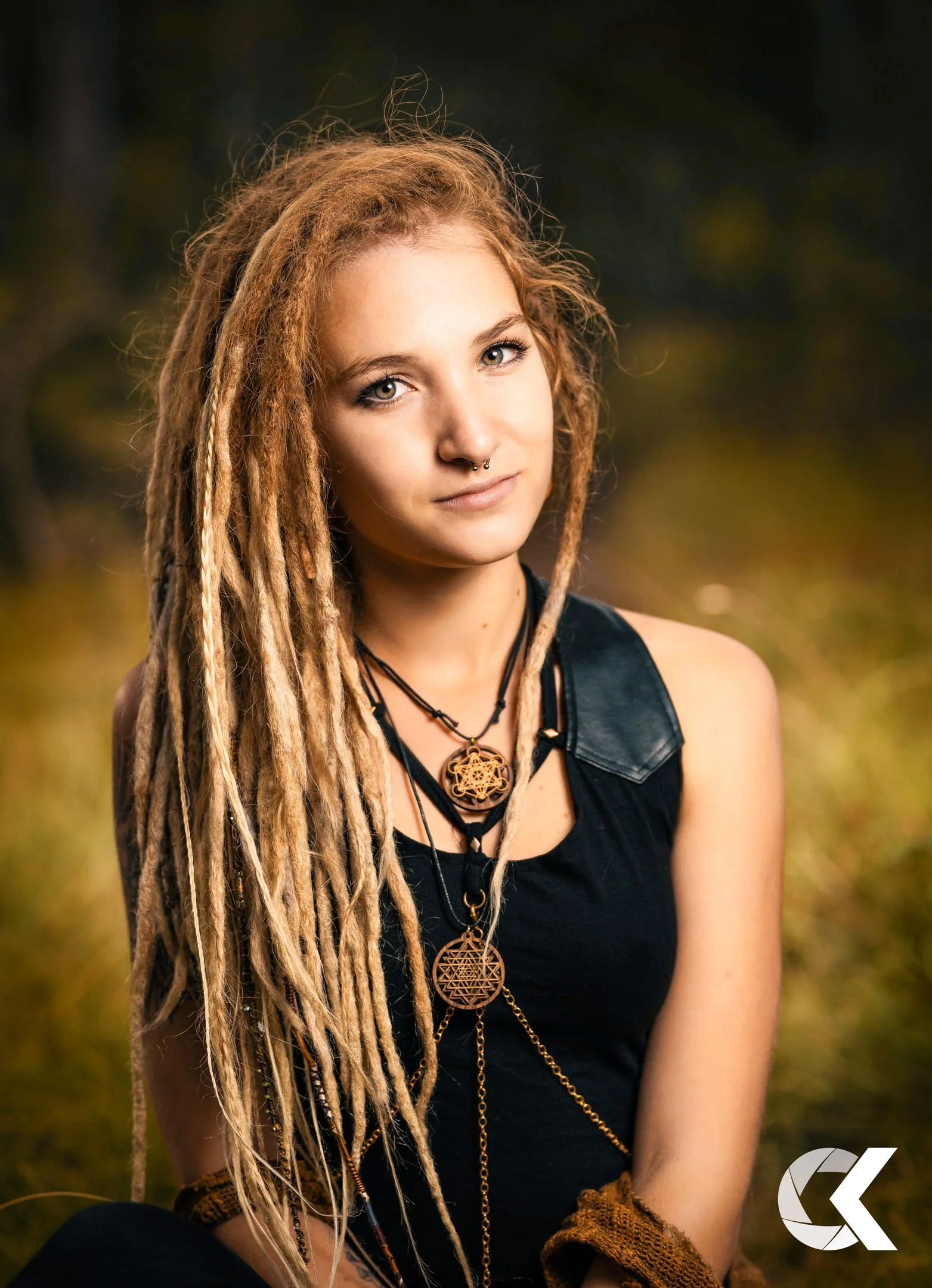 Young woman with long blond dreadlocks, wearing black tank top and layered necklaces with intricate pendants, sitting outdoors in a natural setting.