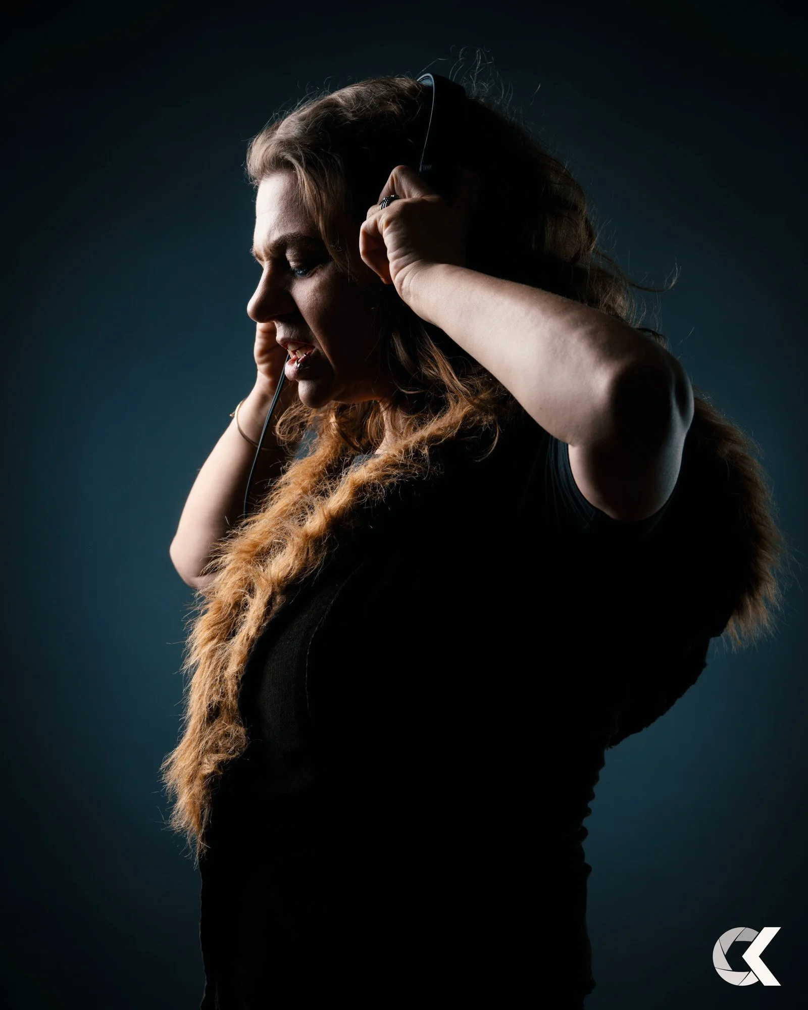 A woman with curly hair wearing headphones, closing her eyes with a concentrated expression against a dark background.