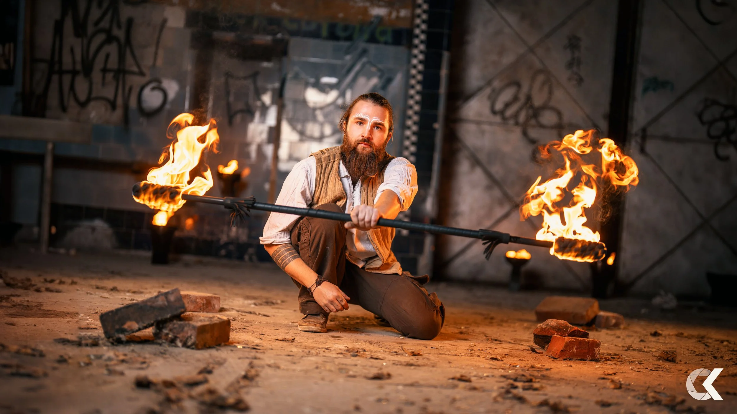 A man with a beard and long hair crouches on the ground holding a fire staff with flames at both ends in an industrial, graffiti-covered setting.