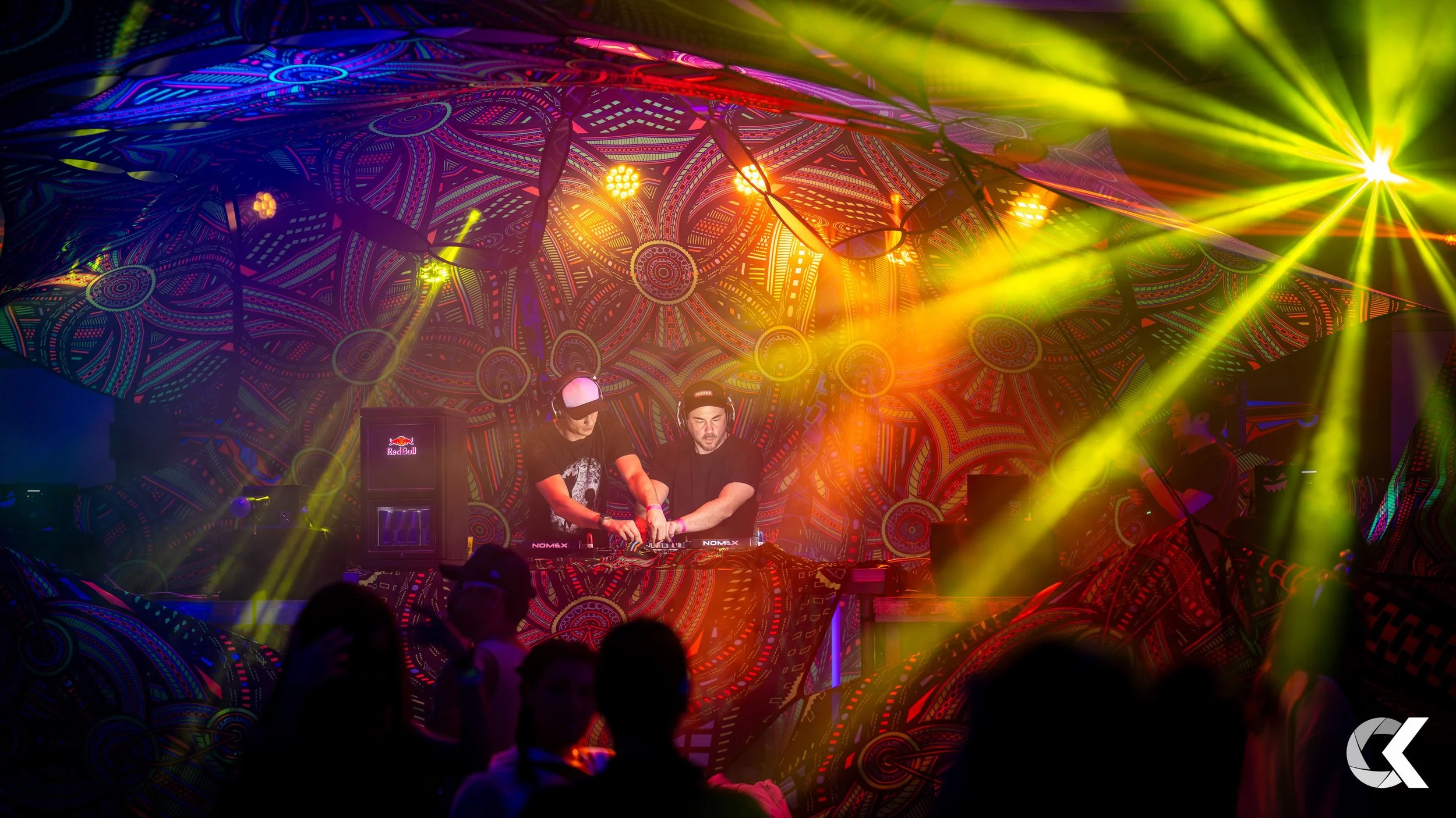 A nightclub scene with two DJs performing at a colorful, patterned stage with yellow and orange lighting and some silhouettes of people in the foreground.