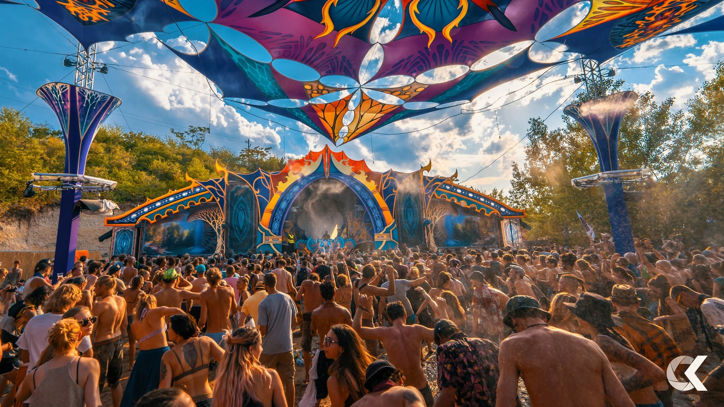 Crowd of people dancing and enjoying an outdoor music festival with a colorful stage and decorative canopy overhead, in a natural setting with trees and a partly cloudy sky.