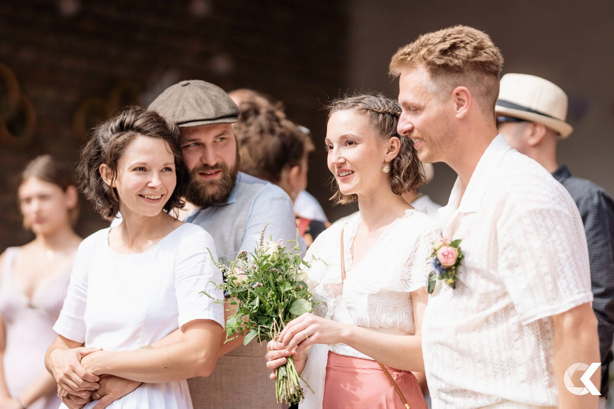 Four people dressed in wedding attire standing together and smiling at a wedding reception, with a woman holding a bouquet of flowers.