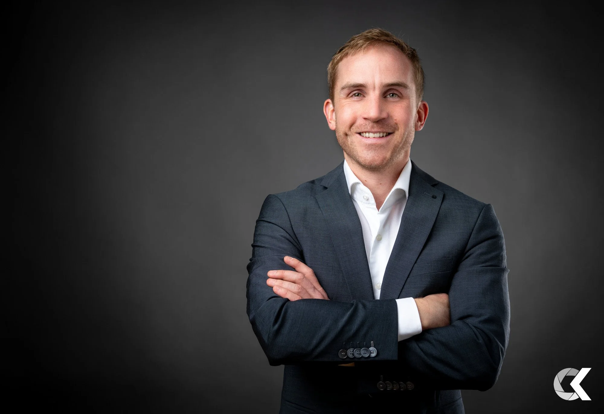 A smiling man in a dark suit with arms crossed, standing against a dark gray background.