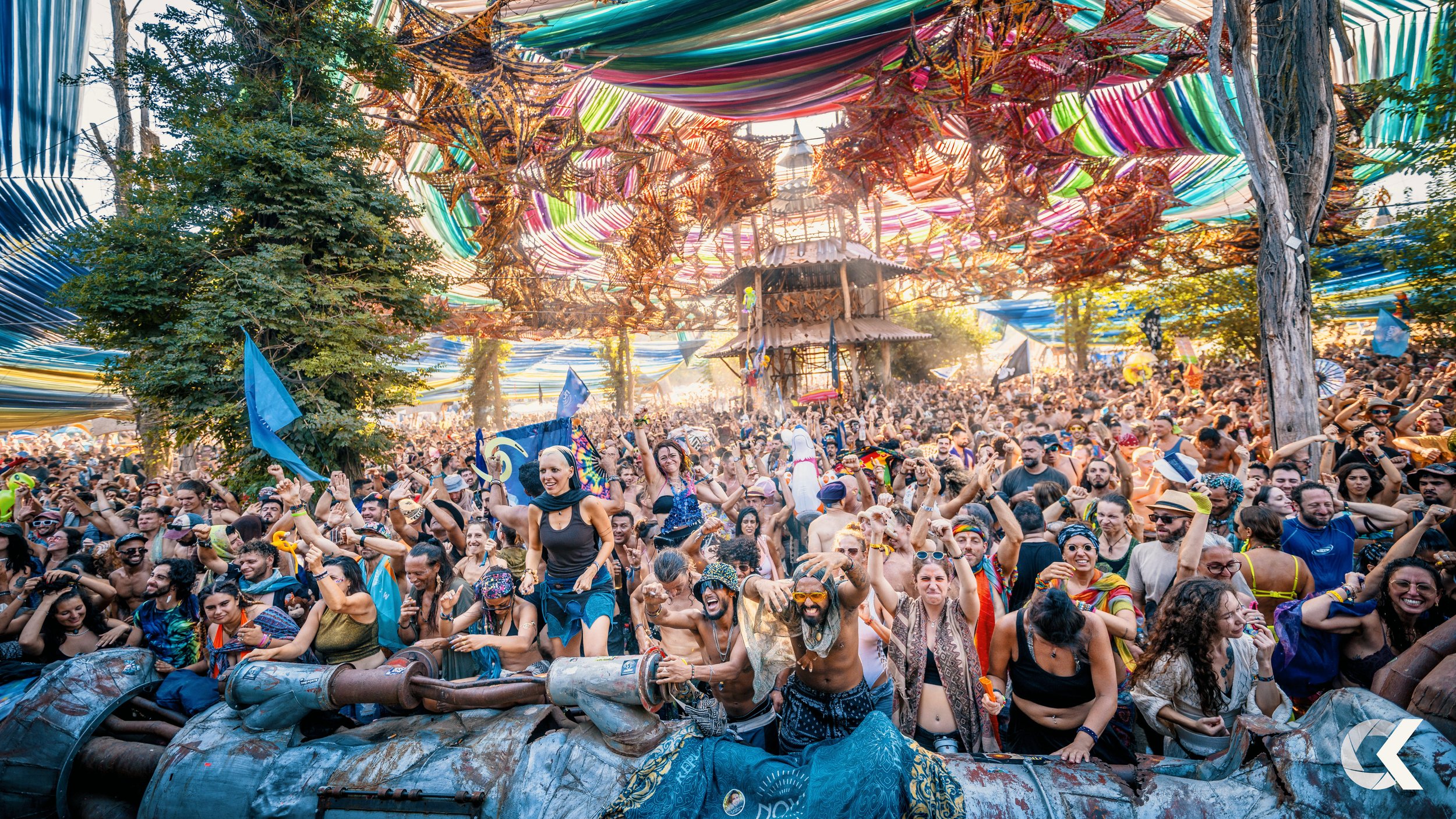 Crowd of people dancing and having fun at an outdoor music festival with colorful fabric decorations and trees in the background.
