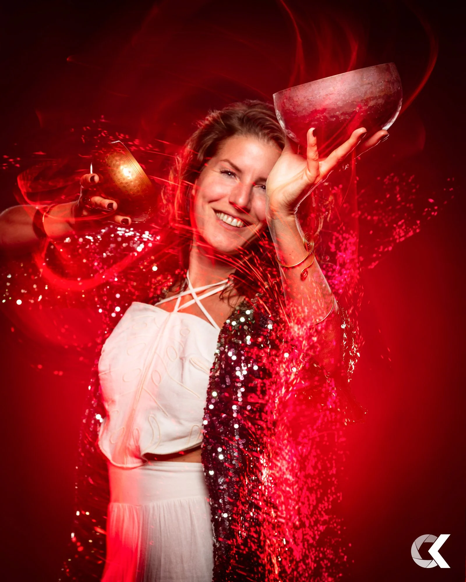 A woman smiling, holding a metallic bowl in one hand and a smaller bowl in the other, surrounded by red sparkles and light effects, wearing a shiny red jacket and white dress.