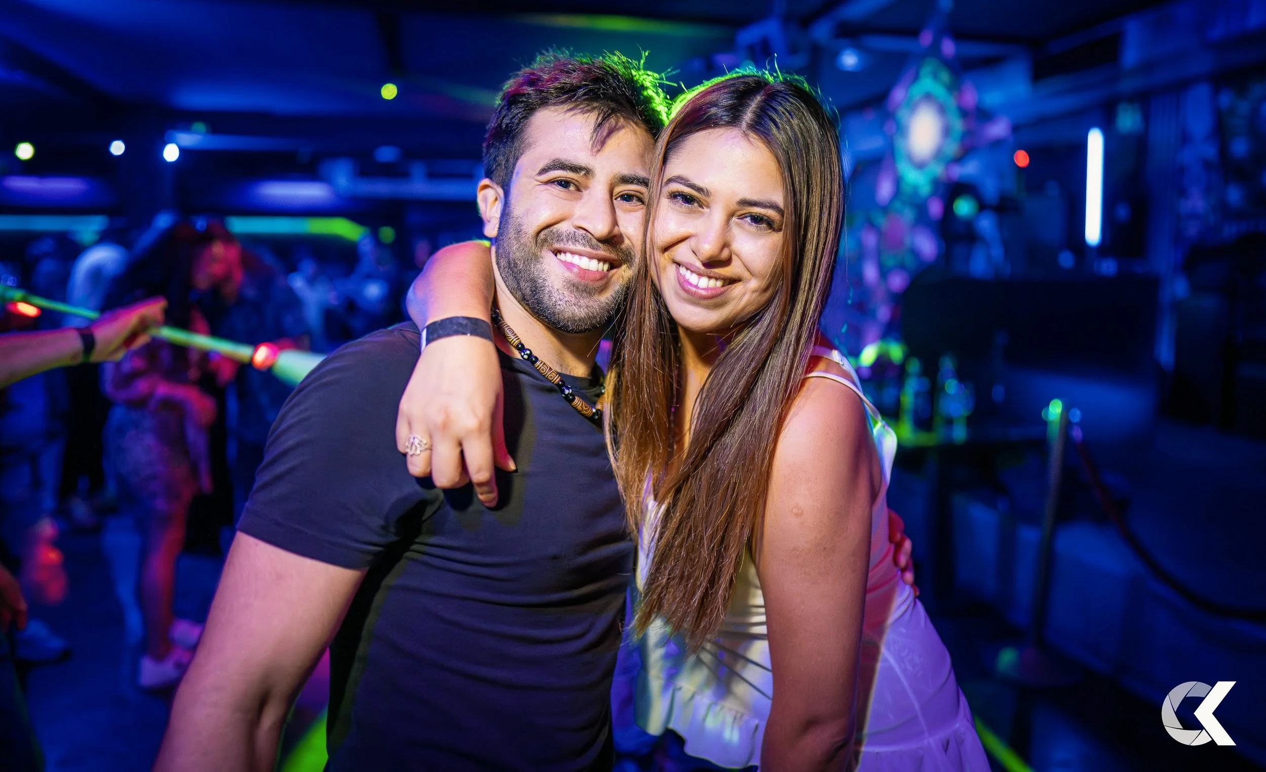 A smiling man and woman with arms around each other in a nightclub, with colorful lights and other people dancing in the background.