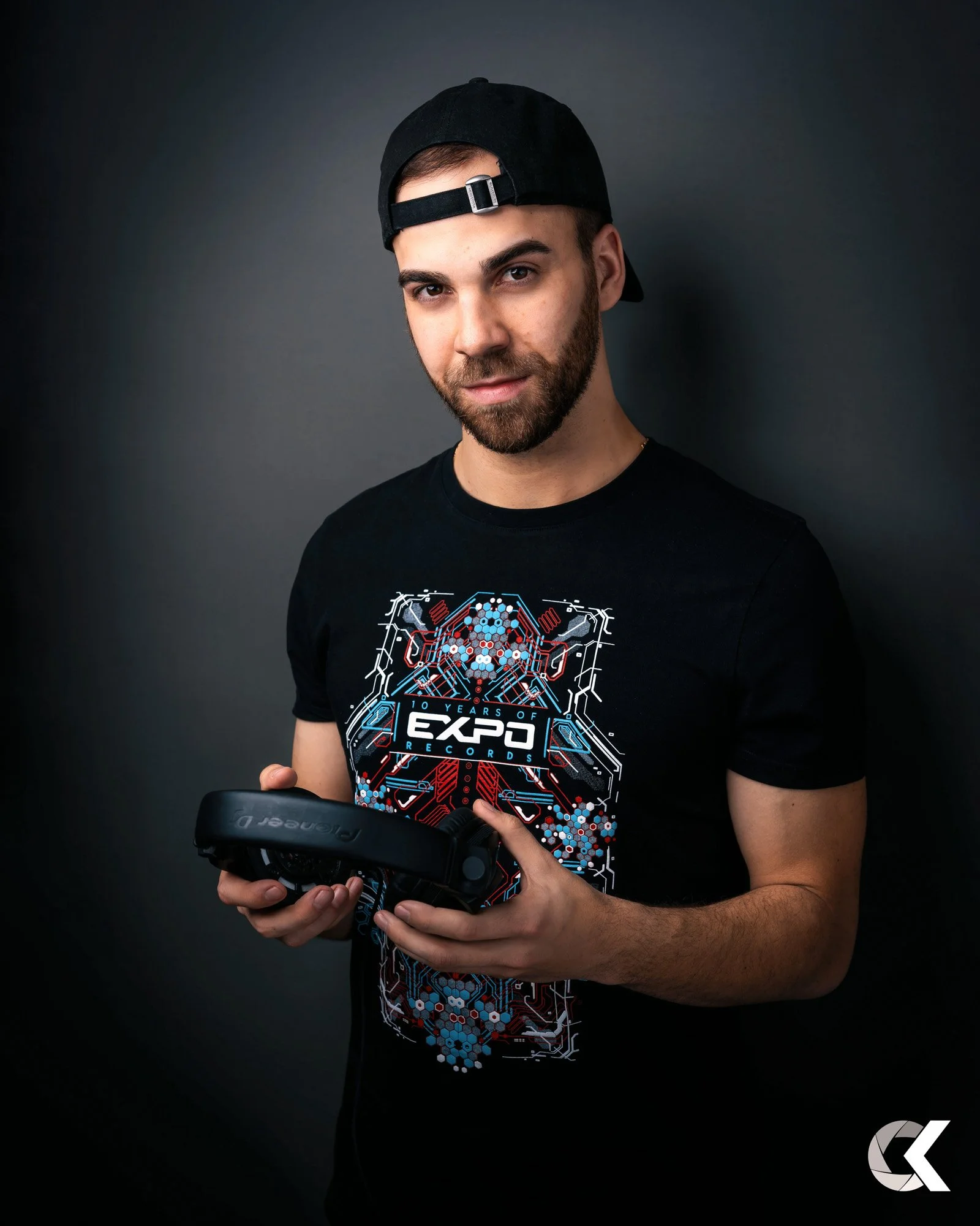 Young man with a beard wearing a black cap backwards and a black t-shirt with electronic circuit graphics, holding a virtual reality headset, standing against a dark gray background.