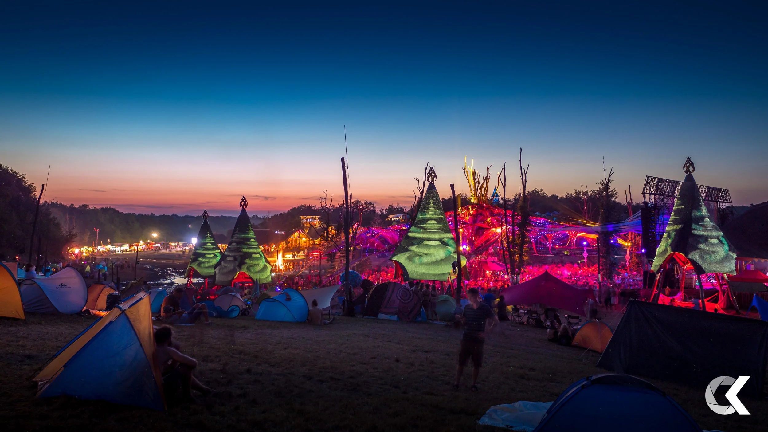 A festival at sunset with tents in the foreground, illuminated trees, and a brightly lit stage with colorful lights in the background.