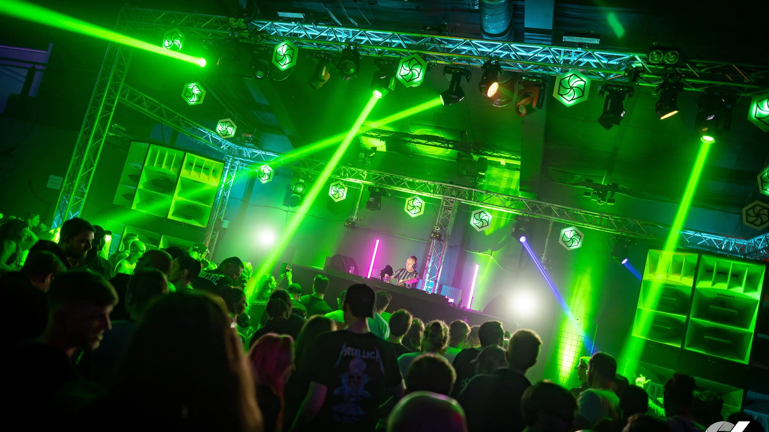 Lively indoor concert scene with a DJ performing on stage, colorful green and purple laser lights, and a crowd of people enjoying the music.