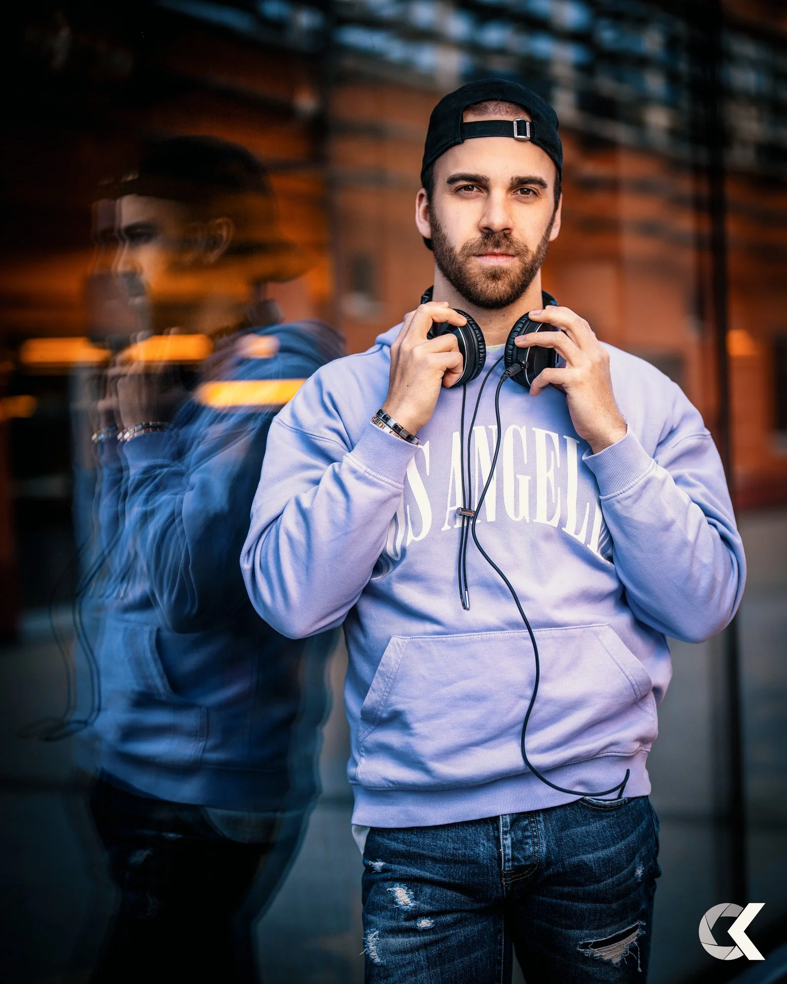 A young man with a beard and dark hair wearing a black backward cap, a lavender hoodie with 'LOS ANGELES' written in white, and ripped jeans. He is holding a pair of headphones around his neck and standing outside with a reflective glass window behin