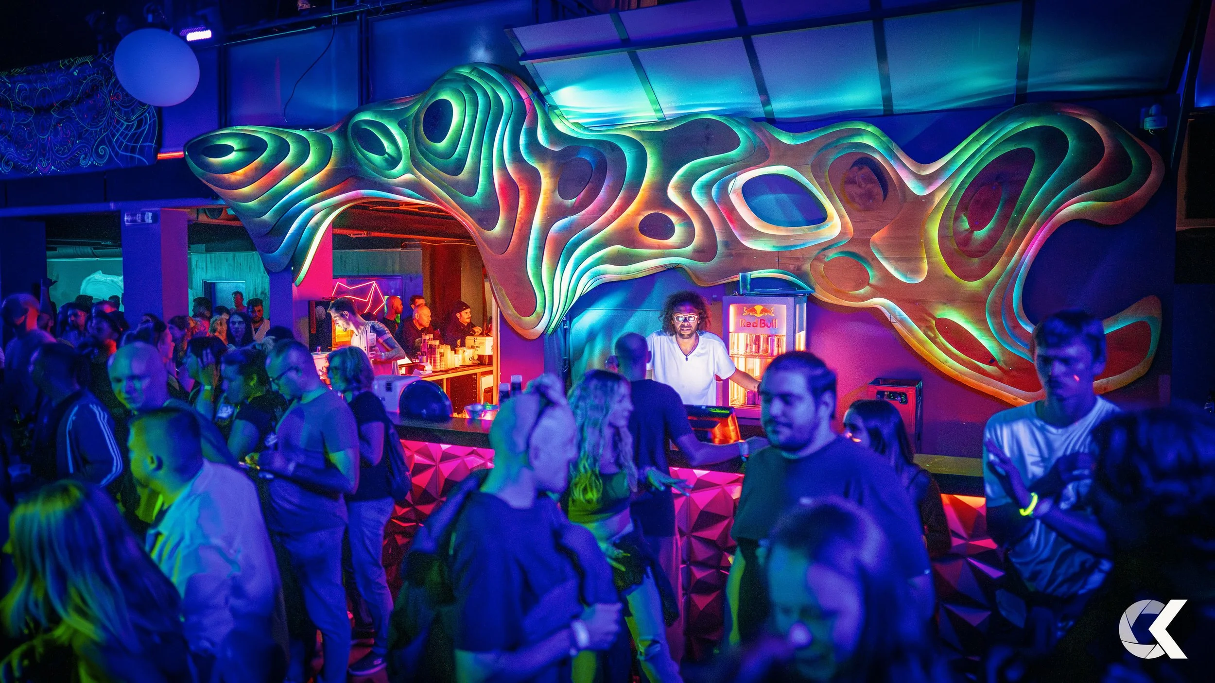 People dancing and socializing at a nightclub with colorful, abstract neon wall art and a bar in the background.