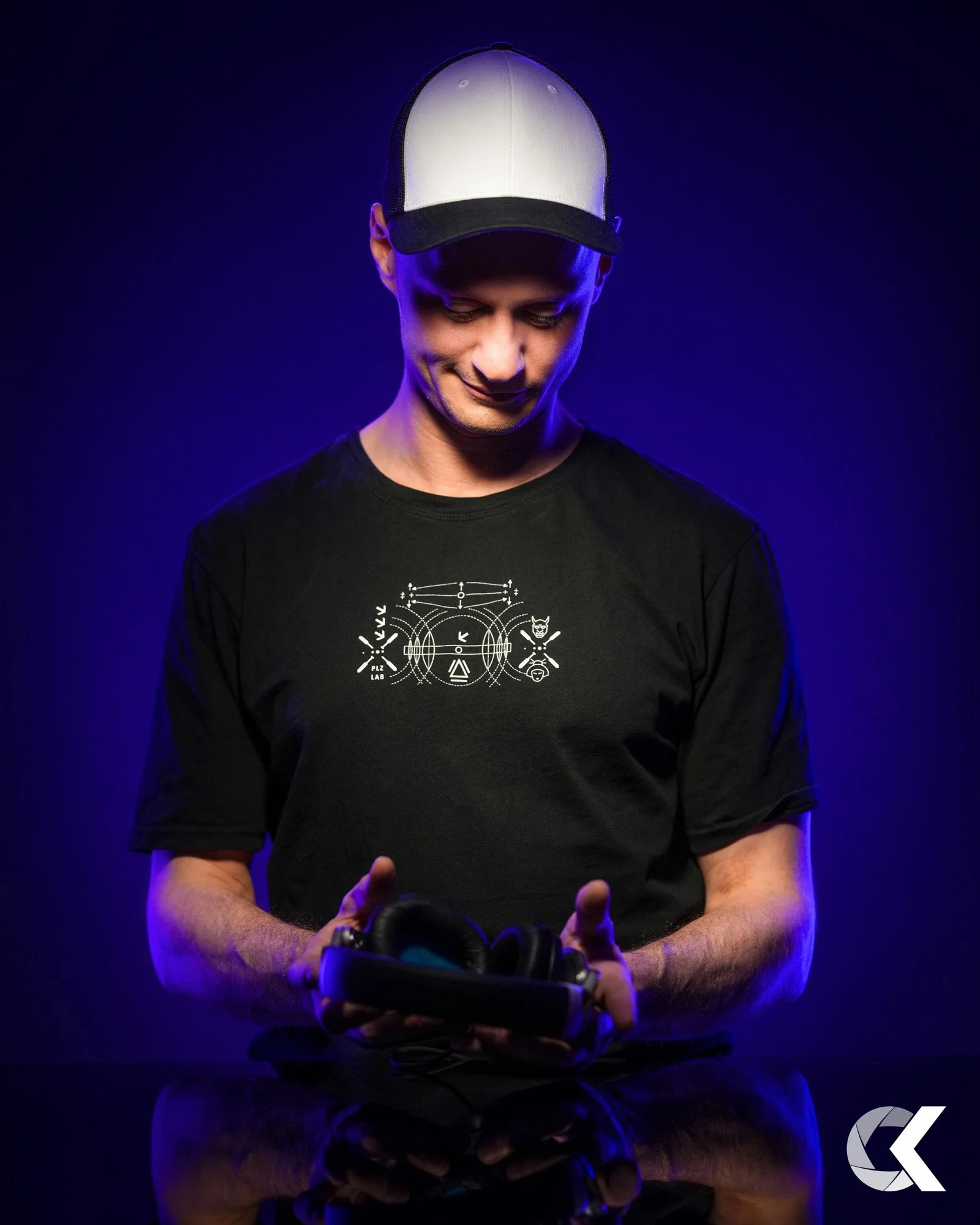 A young man wearing a black T-shirt and a baseball cap, looking down and holding headphones, with blue lighting and a dark background.