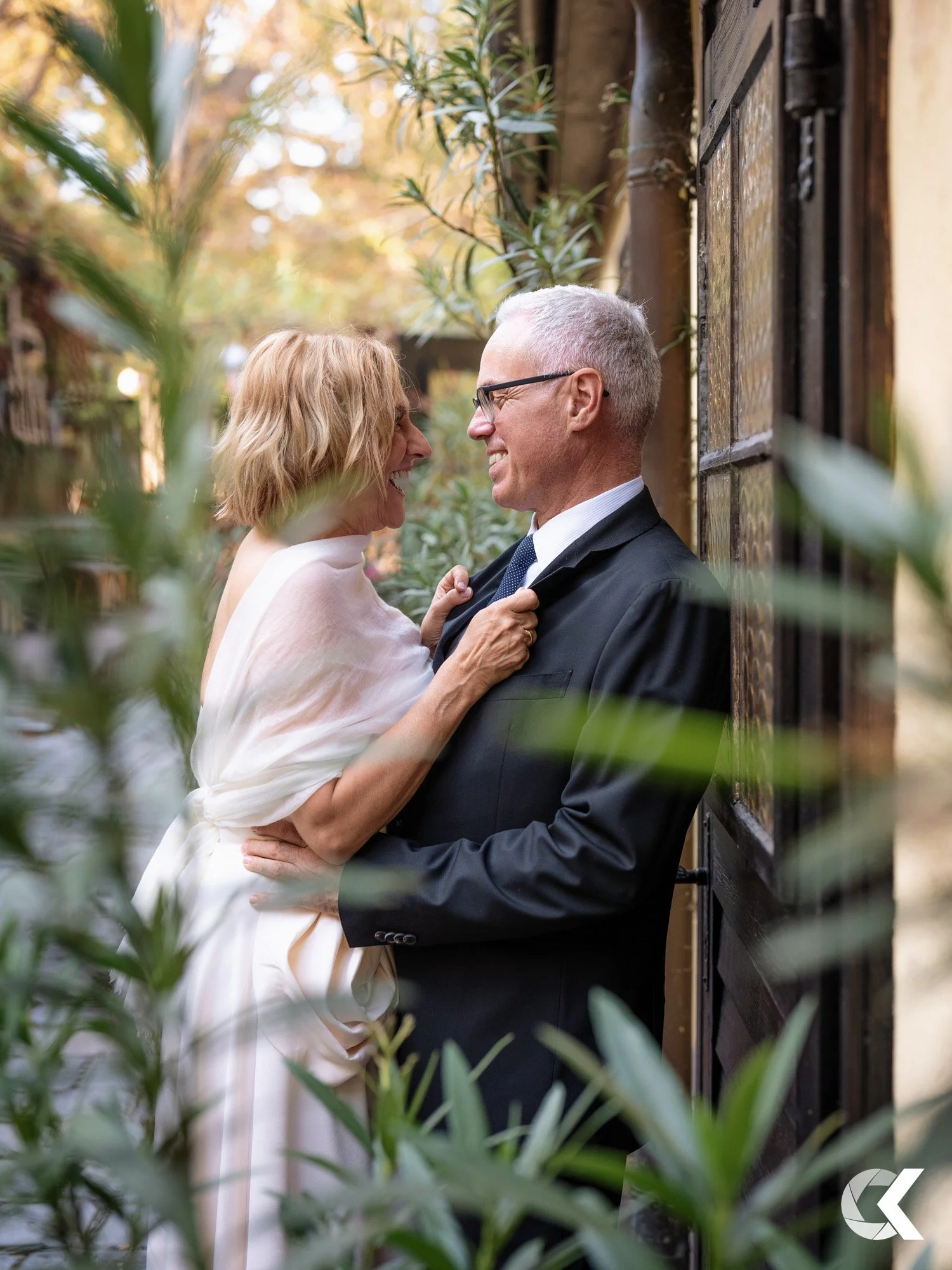 A middle-aged woman and man smiling and looking at each other, standing close with greenery around them, in an outdoor setting.