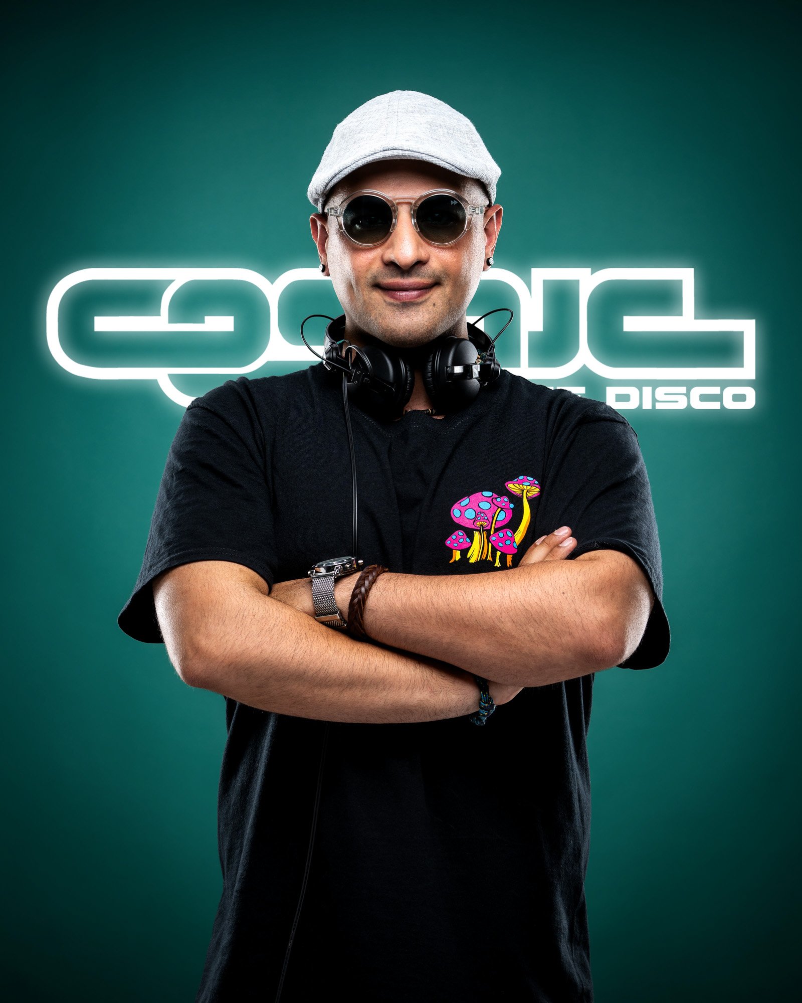 A man wearing sunglasses, a gray cap, and a black t-shirt with a colorful mushroom design, standing with arms crossed in front of a teal background with a neon sign that reads 'COOL DISCO'.