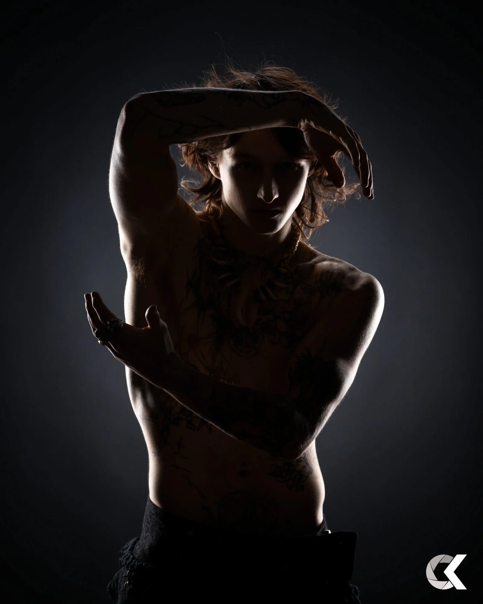 A person with tattoos on their arms and chest, posing with one arm above their head and the other bent across their chest, against a dark background, with dramatic lighting that highlights their silhouette.