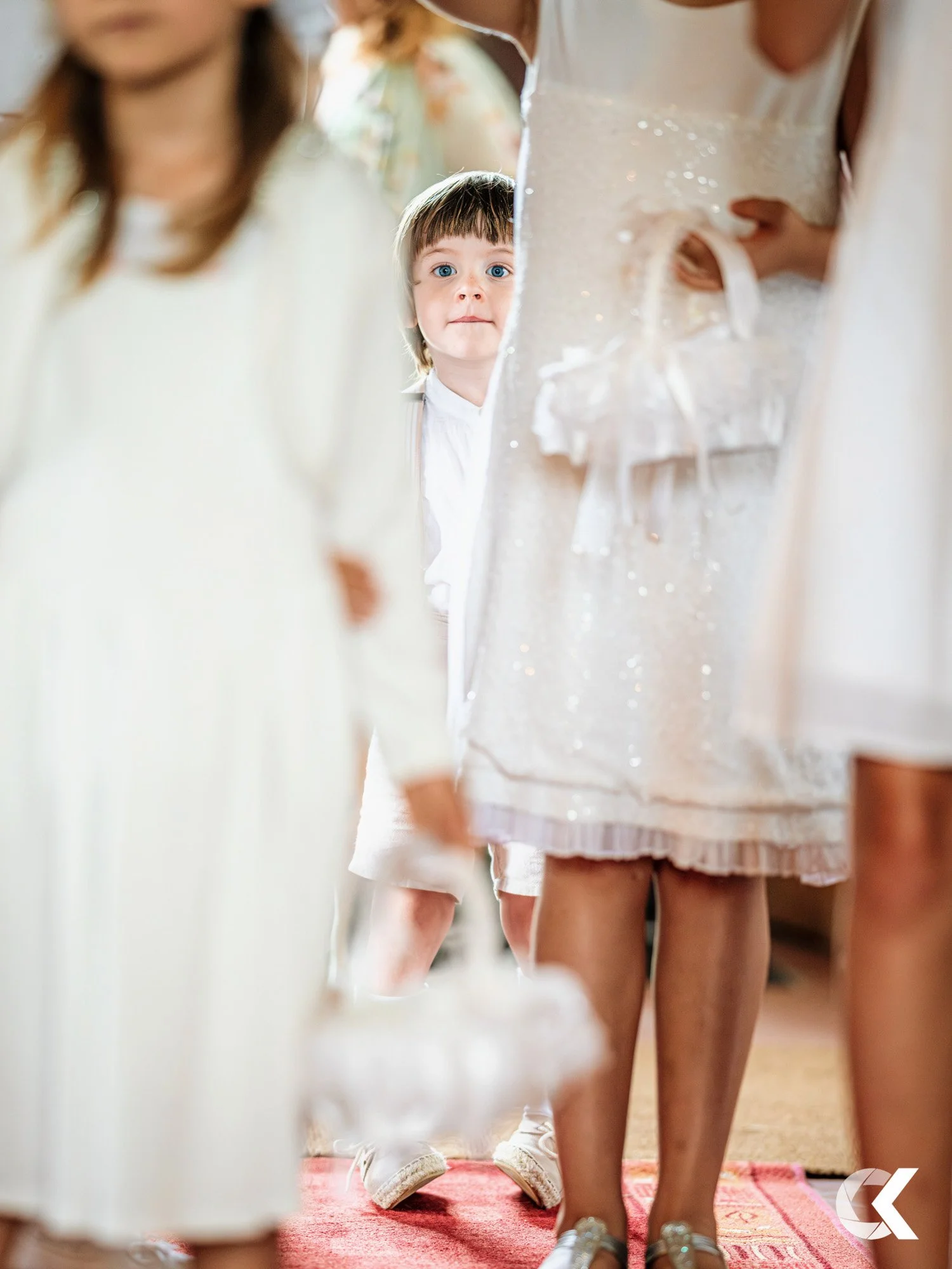 A young boy with blue eyes looks through a gap between women dressed in white, possibly at a wedding or formal event.