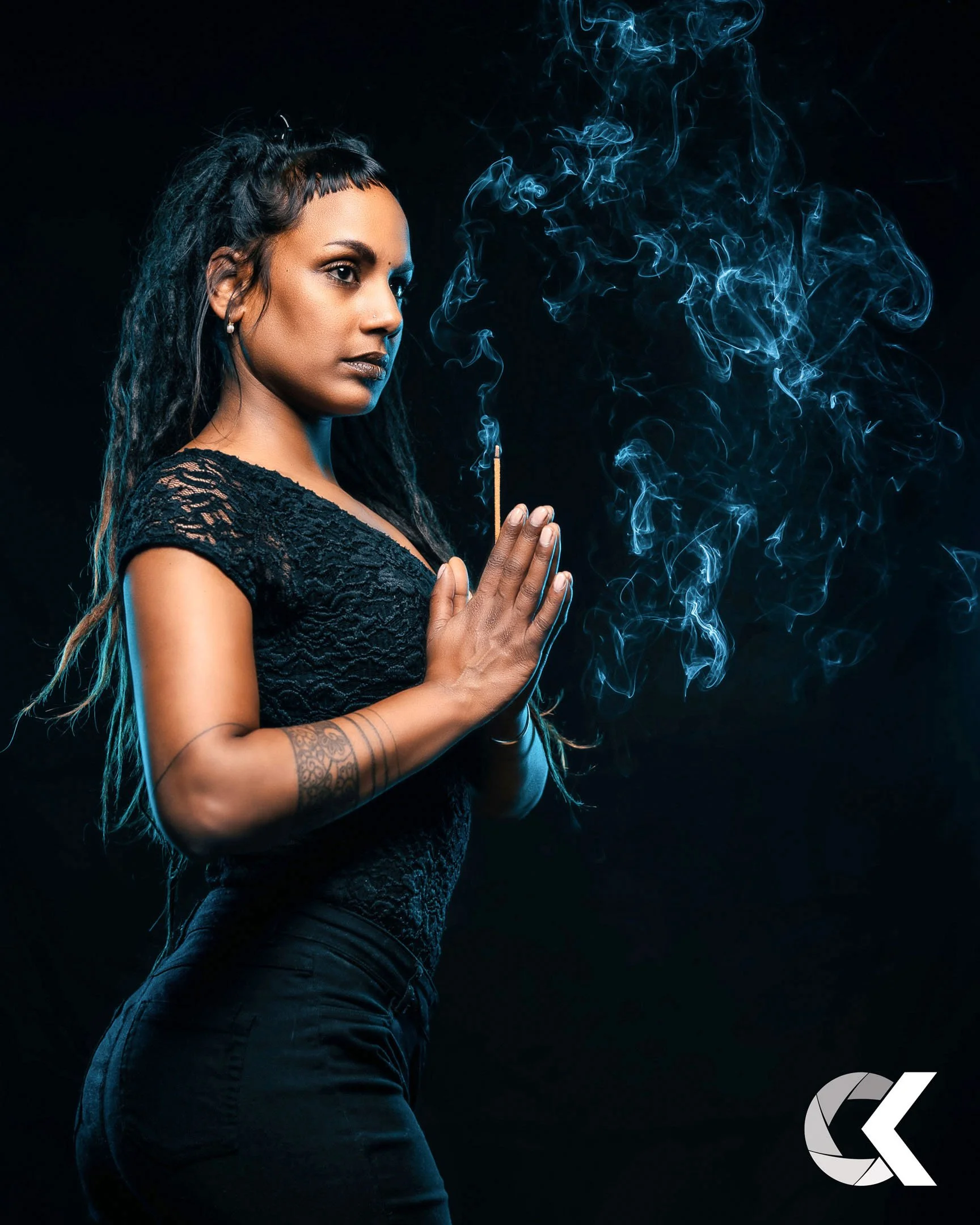 A woman dressed in black with a tattoo on her arm holding a lit matchstick, with blue smoke swirling around her against a dark background.