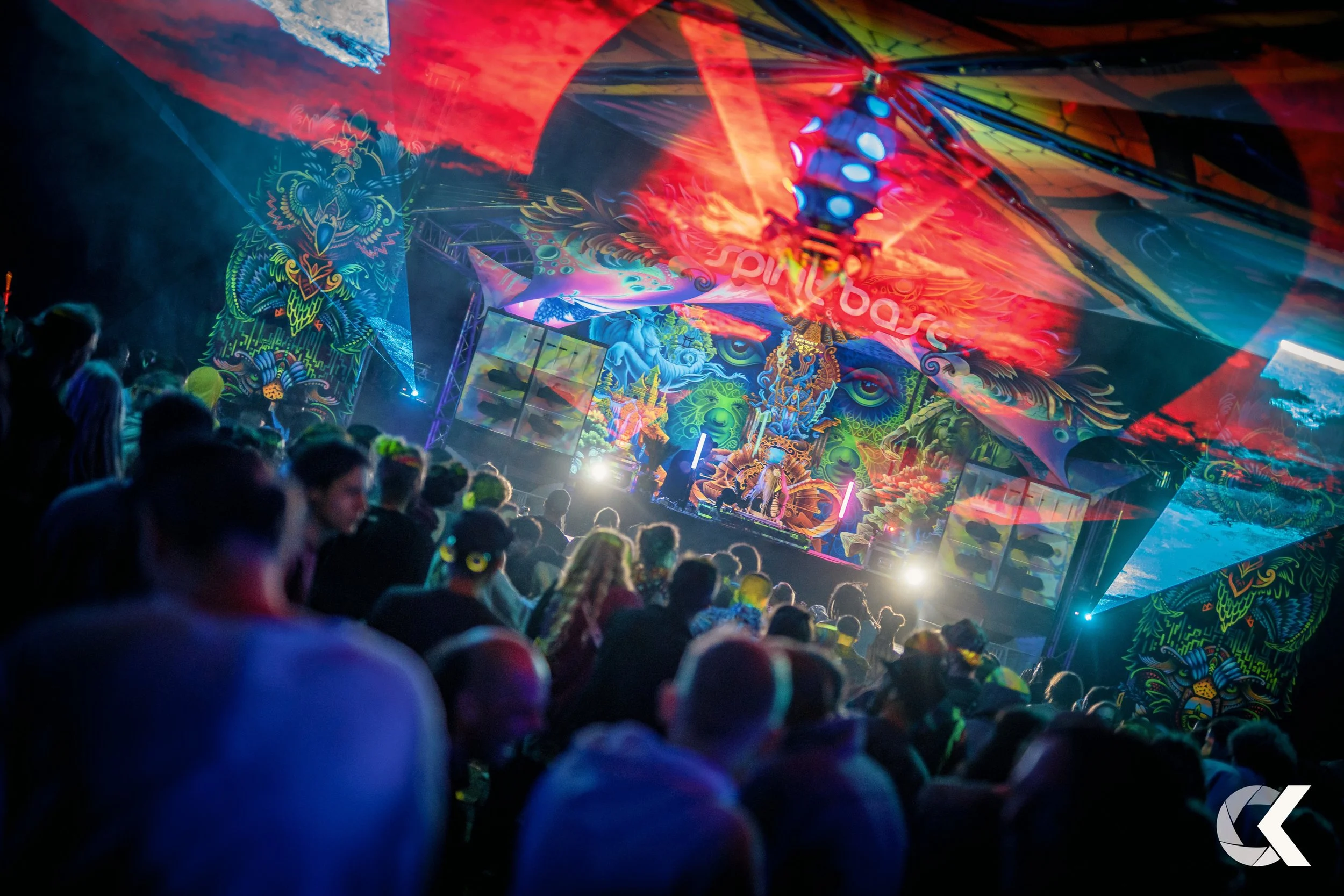 Crowd attending a colorful music festival or concert with vibrant neon and LED lights, psychedelic artwork, and a stage featuring a DJ.
