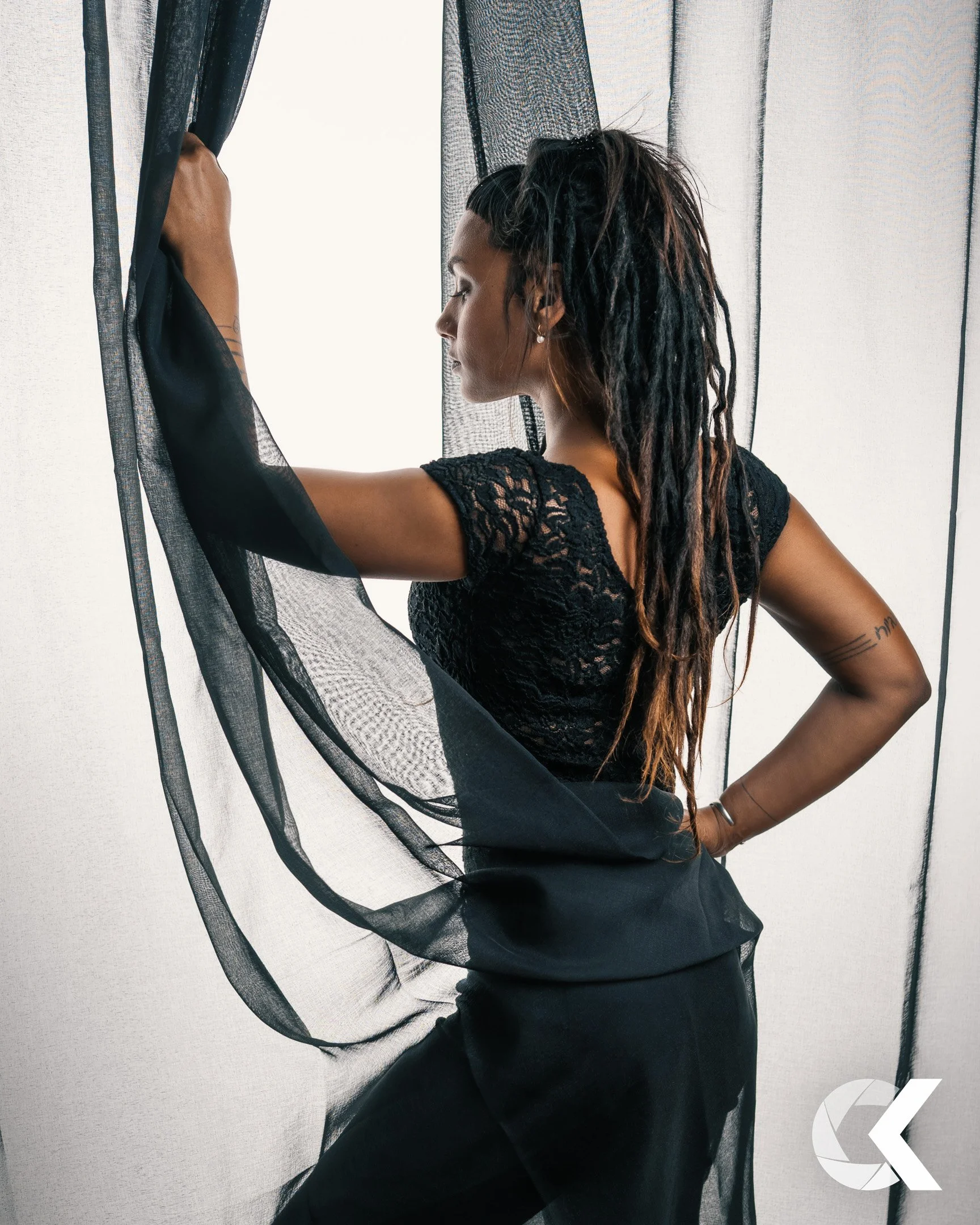 A woman with dreadlocks wearing a black lace top and black pants, standing near a window with sheer curtains, holding one curtain back and looking out.
