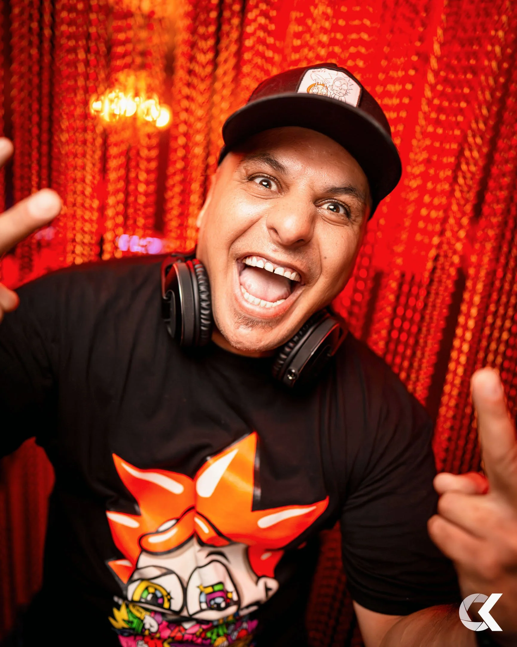 A man smiling and making a peace sign with his hand, wearing a black T-shirt with anime characters, a black cap, and headphones around his neck, against a red curtain background.