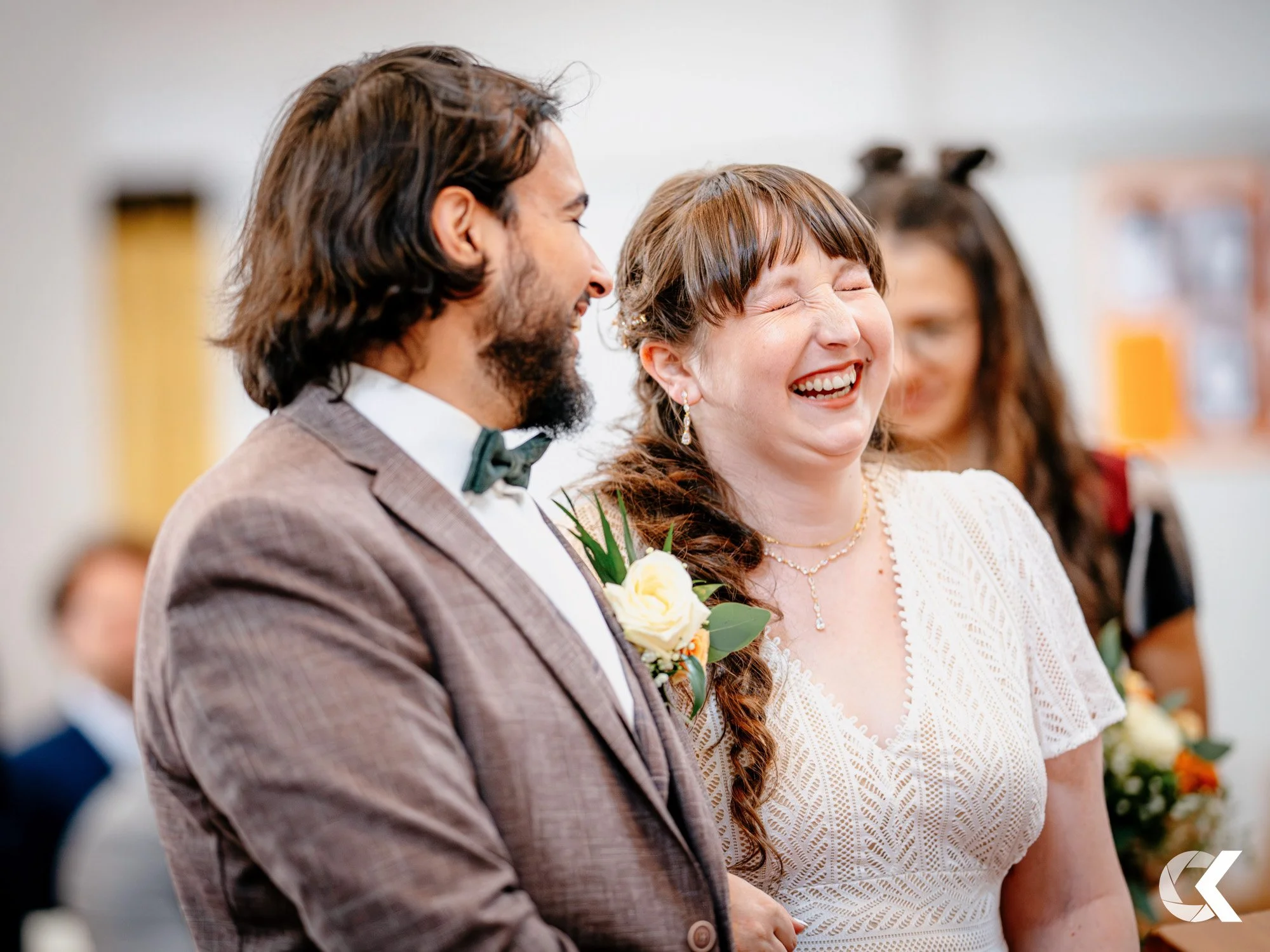 A joyful woman and a man at a wedding ceremony, both smiling and laughing, with a woman in the background holding flowers.