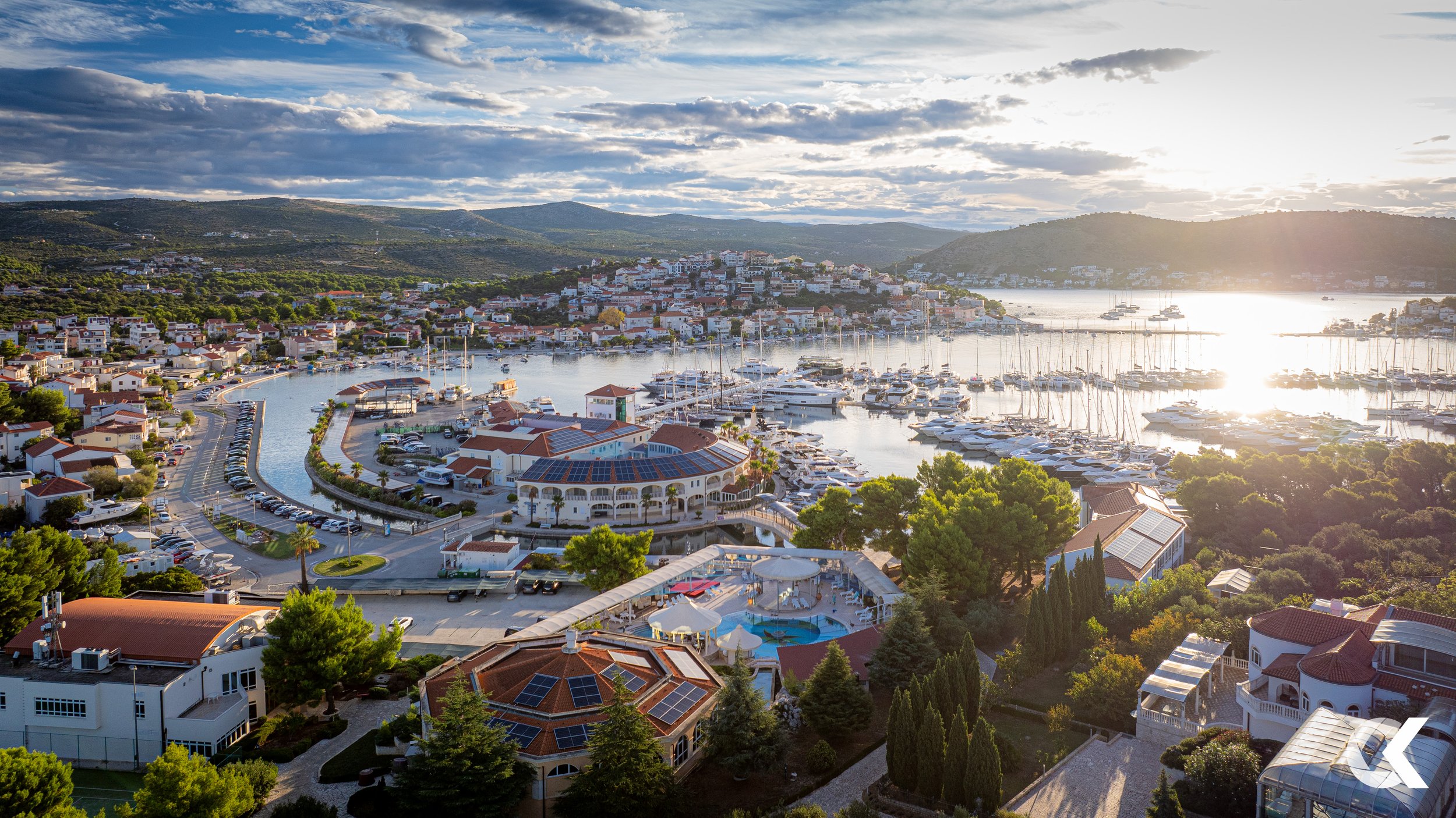 A scenic view of a marina with numerous sailboats and yachts docked in calm water, surrounded by houses and green trees, with hills in the background and the sun setting over the horizon.