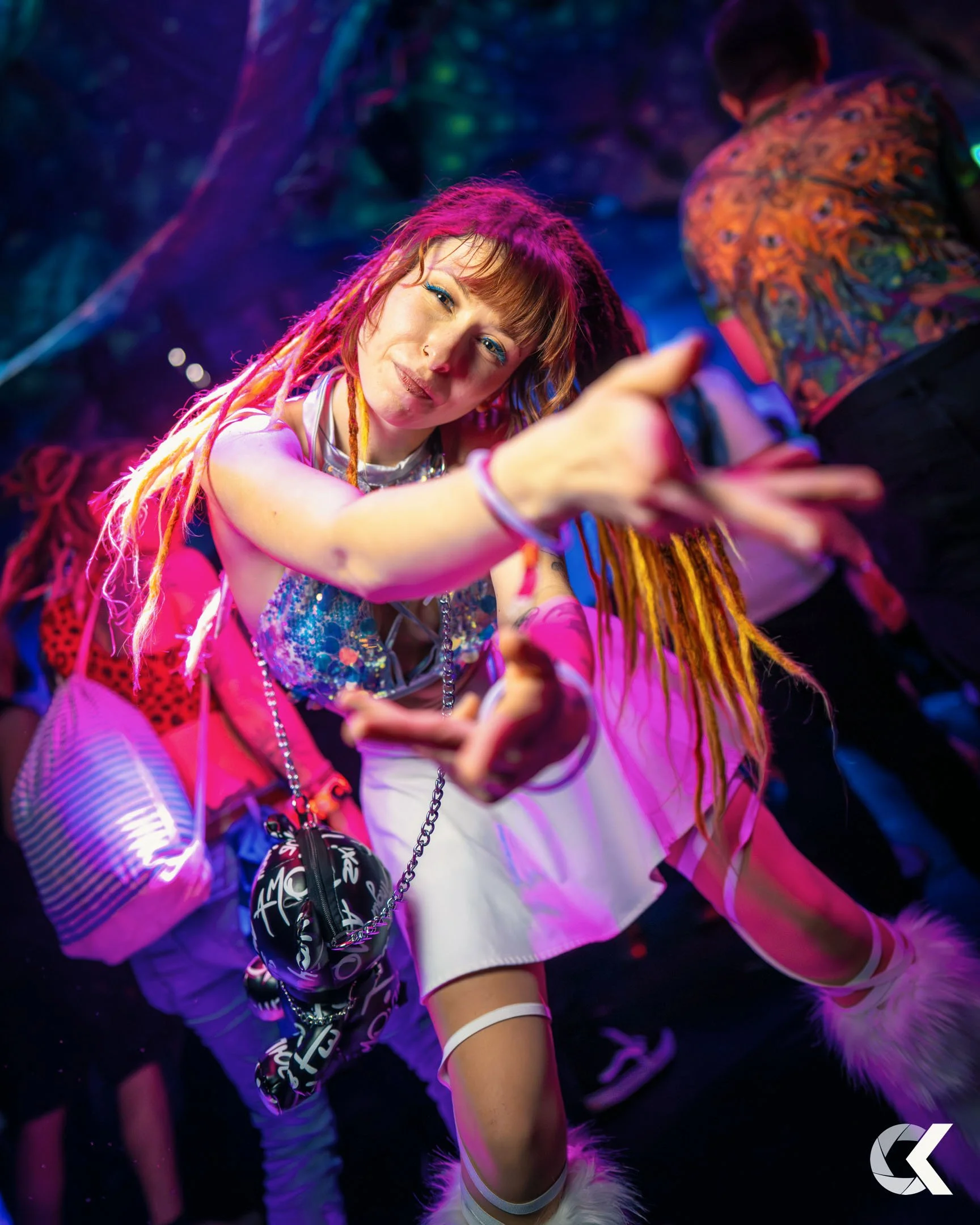 A woman with colorful dreadlocks and vibrant makeup reaching toward the camera at a vibrant party or club.