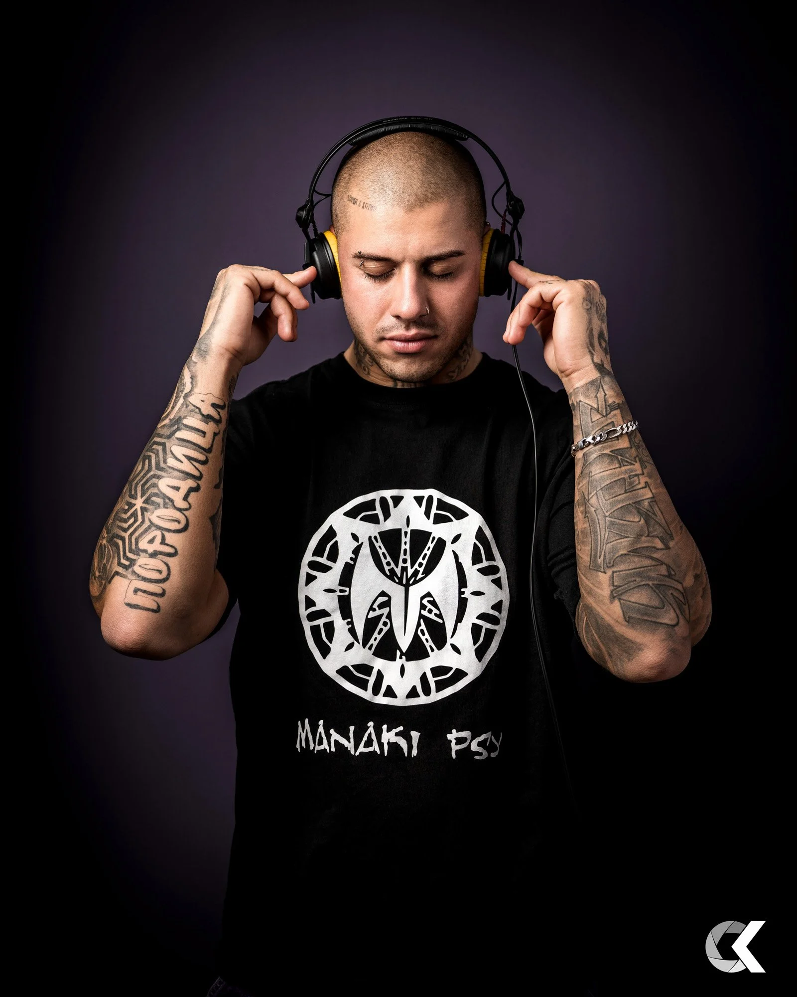A man with tattoos on his arms and a shaved head wearing a black t-shirt with a white graphic and the words 'Manaki Psy', listening to music through large over-ear headphones with closed eyes.