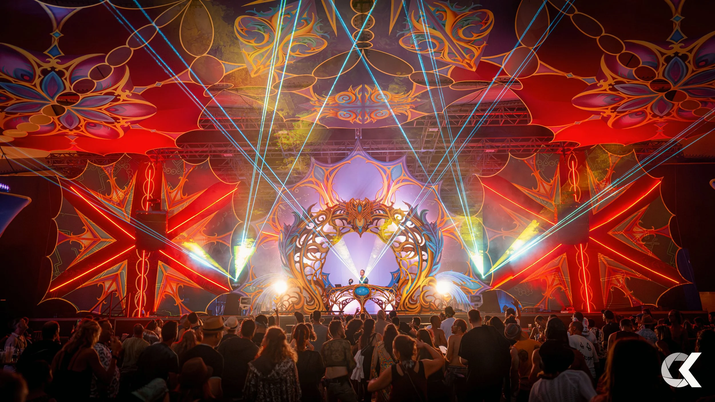 A large electronic dance music event with vibrant lighting, laser beams, and intricate stage designs. The crowd is watching a DJ perform on an elaborate stage with colorful, ornate decorations.