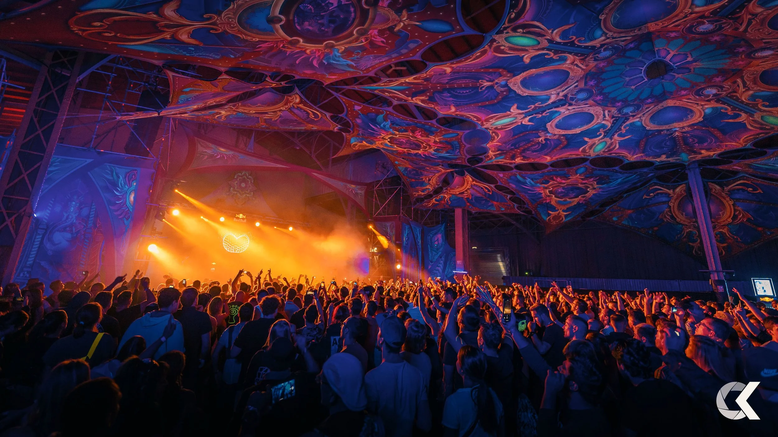 Crowd of people at a concert or music festival with colorful, psychedelic ceiling and stage lighting.