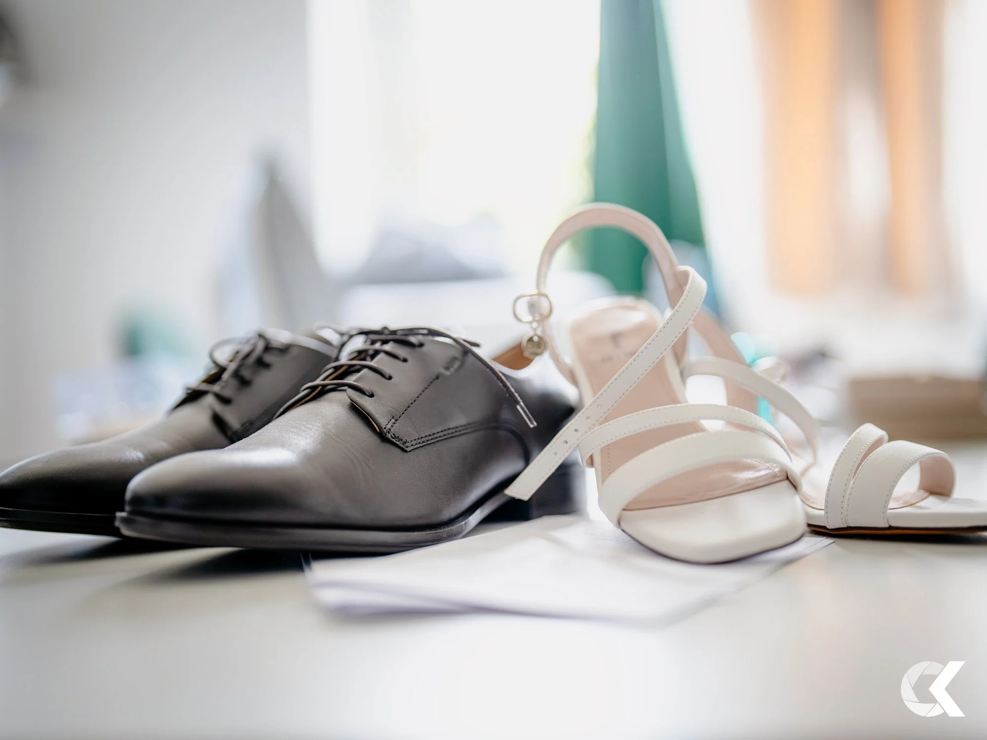 Black formal shoes and white high-heeled sandals on a surface, with a blurred background.