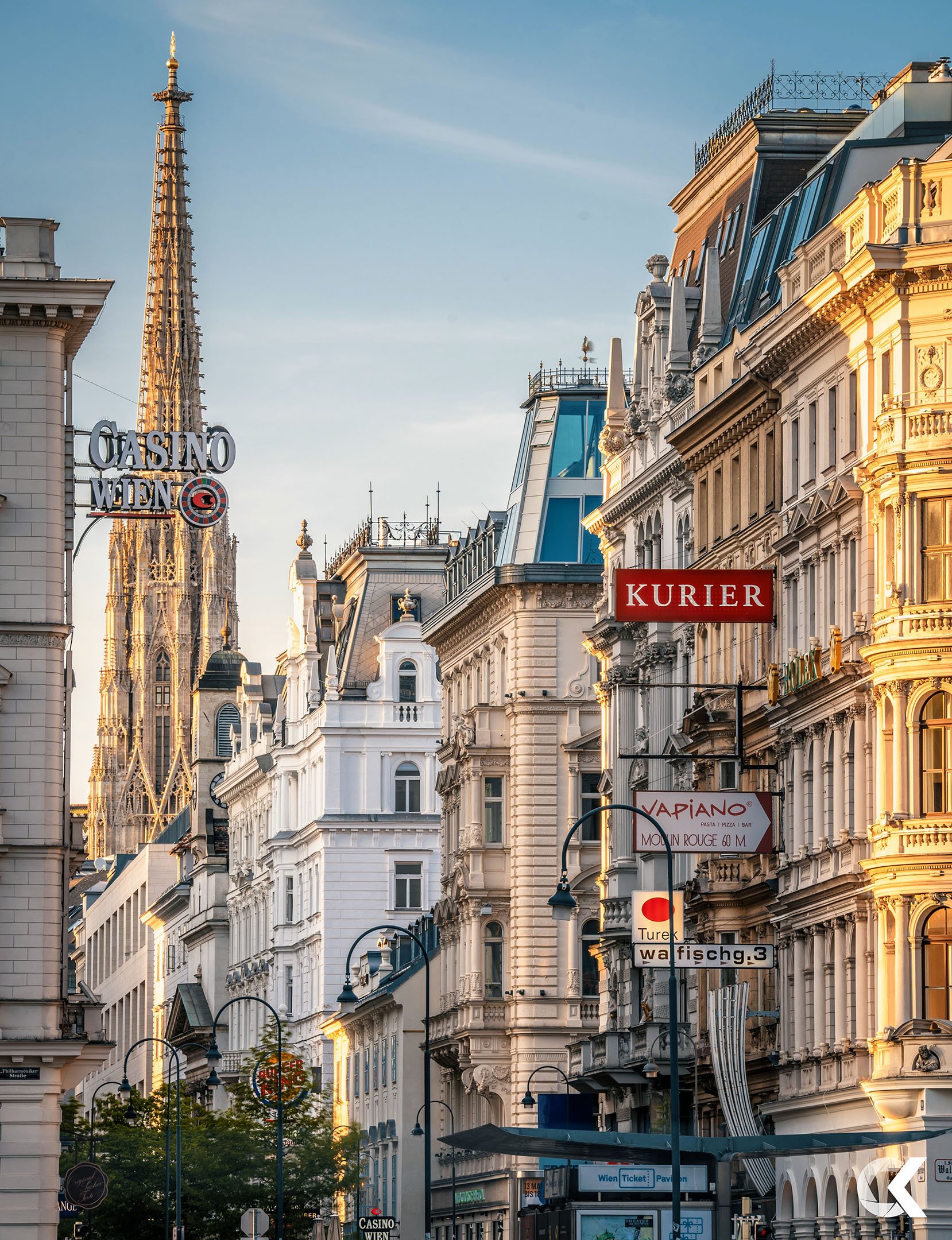 Street view of historic European buildings with signs, including 'Kurier' and 'Vapiano', and the spire of St. Stephen's Cathedral in Vienna in the background during sunset.