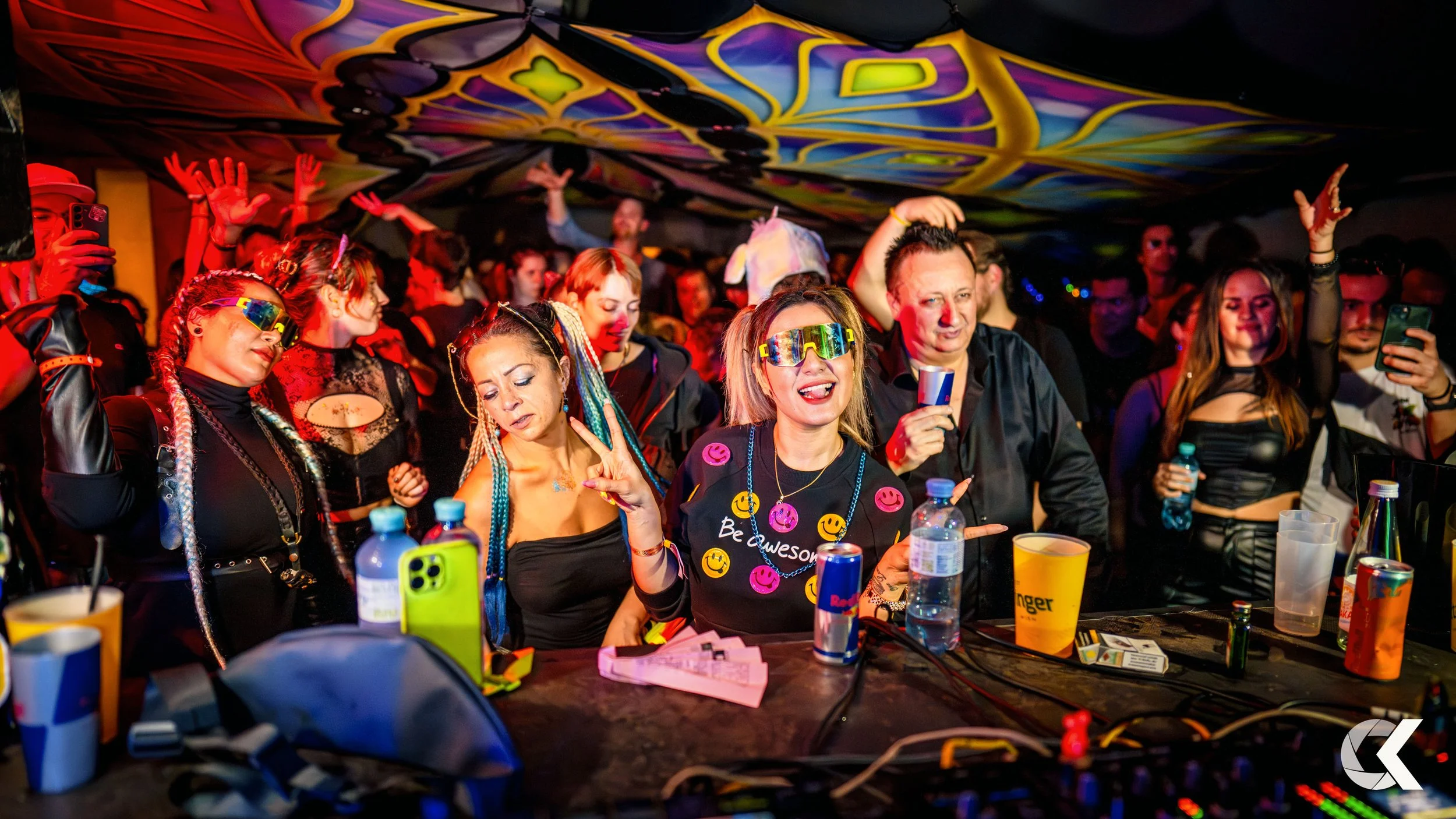 People dancing and enjoying a party with colorful lighting, some wearing futuristic sunglasses, at a crowded indoor event with a decorated ceiling.