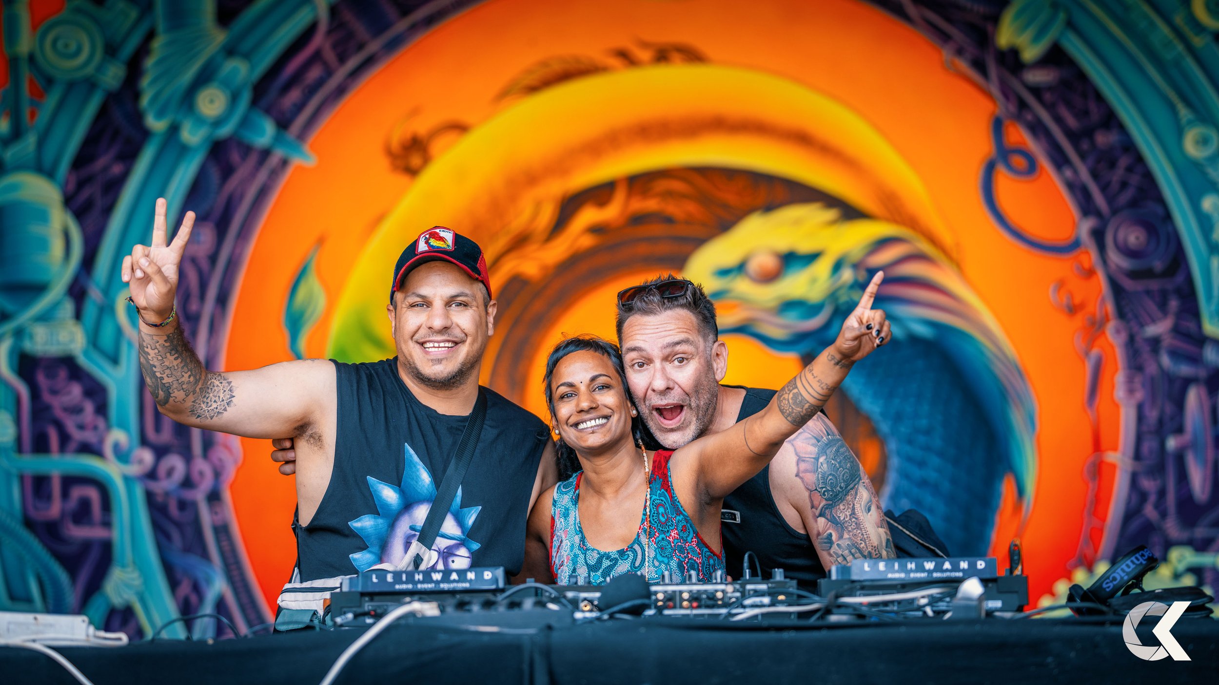 Three festival-goers smiling and dancing behind a DJ booth at an electronic dance music event, with a vibrant, colorful mural of a wave in the background.